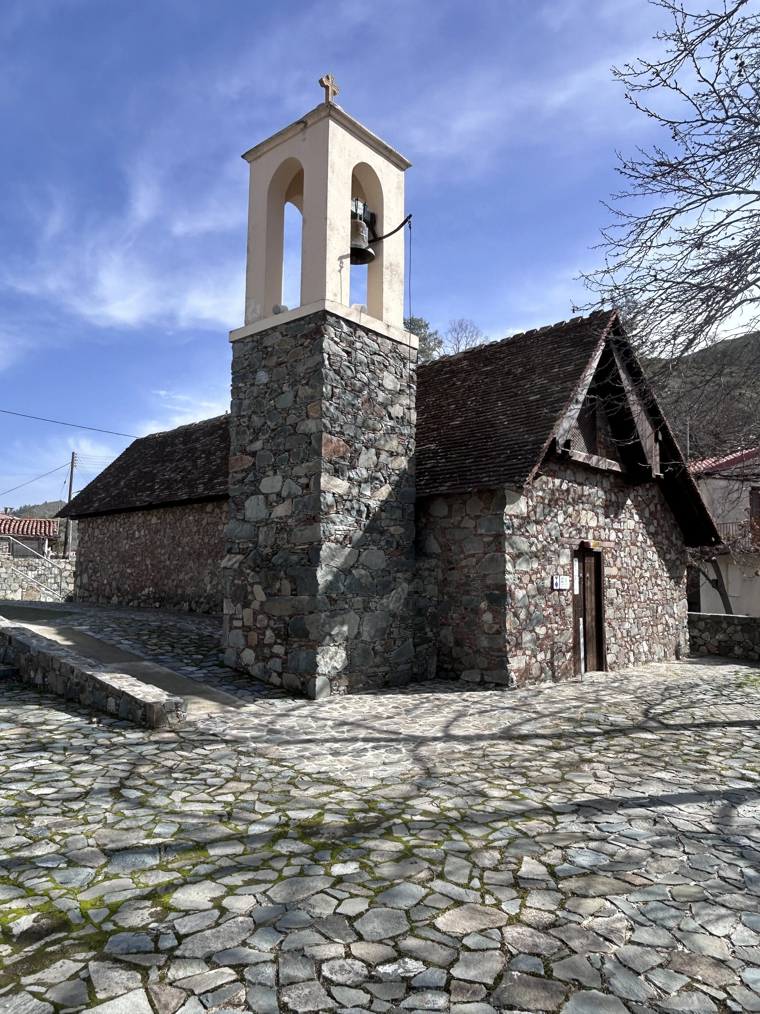 Small stone church with a bell tower and cross on top, set against a blue sky with sparse clouds, with a cobblestone courtyard and leafless trees.