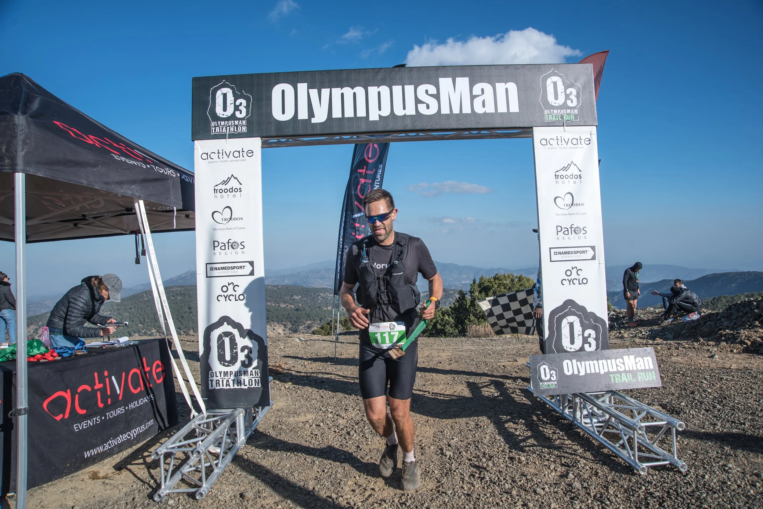 A male trail runner crossing the finish line at the OlympousMan Triathlon, wearing black running gear, sunglasses, and a race bib, with mountains and a blue sky in the background.