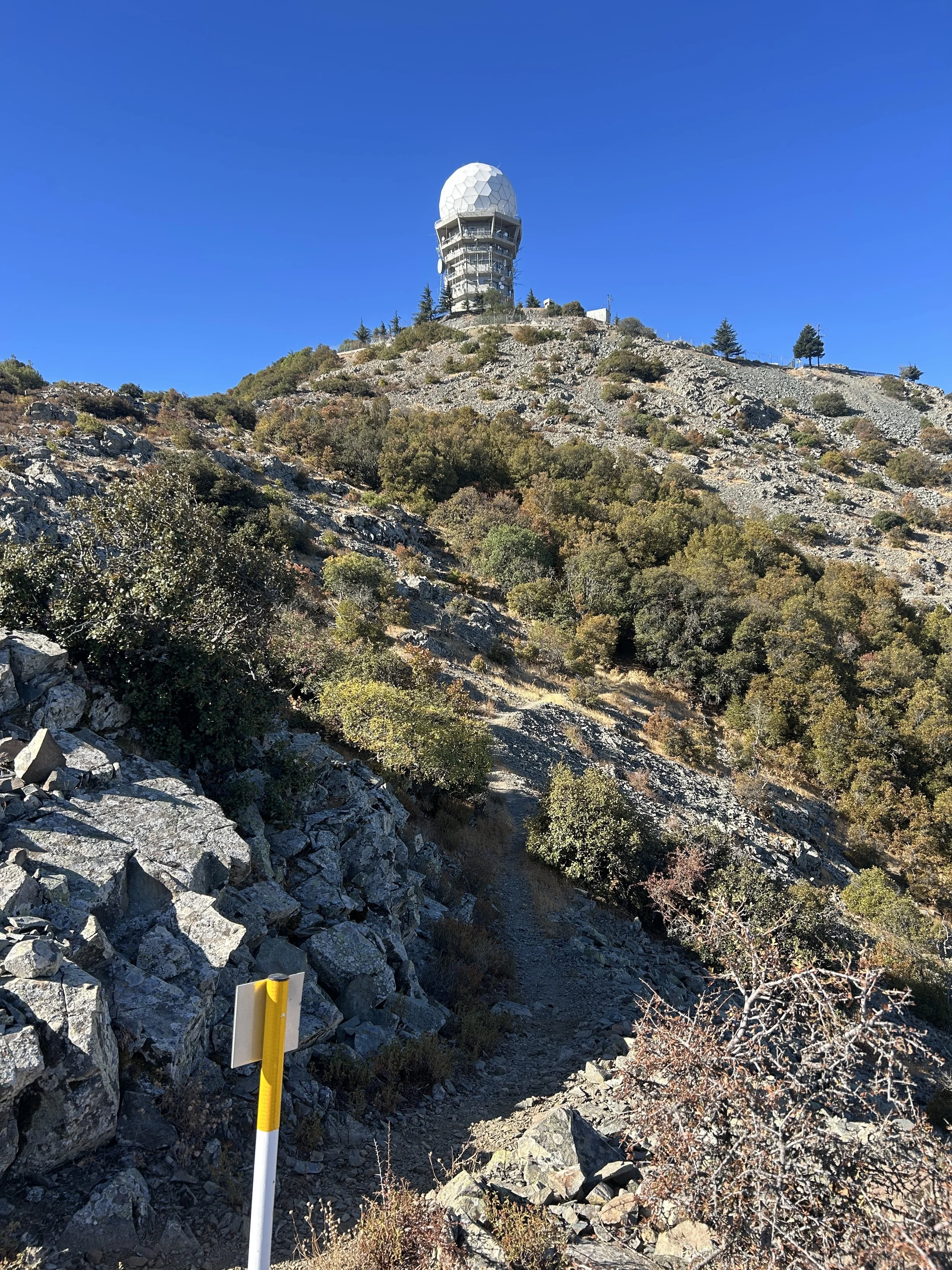 A rugged mountain trail with rocks and sparse vegetation, leading up towards a large radar or communication tower with a white dome at the top, against a clear blue sky.