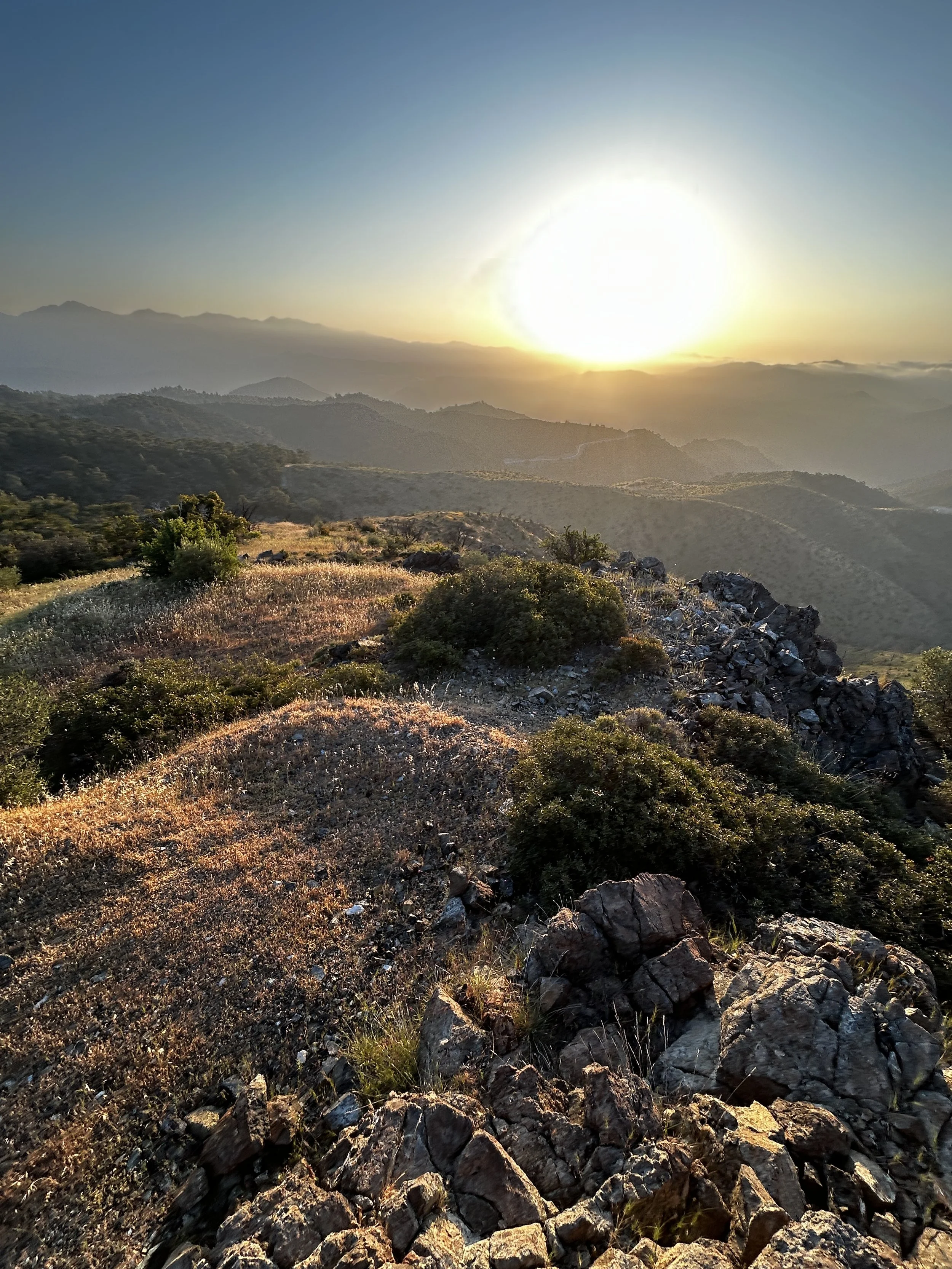 Sunset over a mountainous landscape with rocky and shrub-covered terrain in the foreground, and layered mountain ranges in the distance under a clear sky.