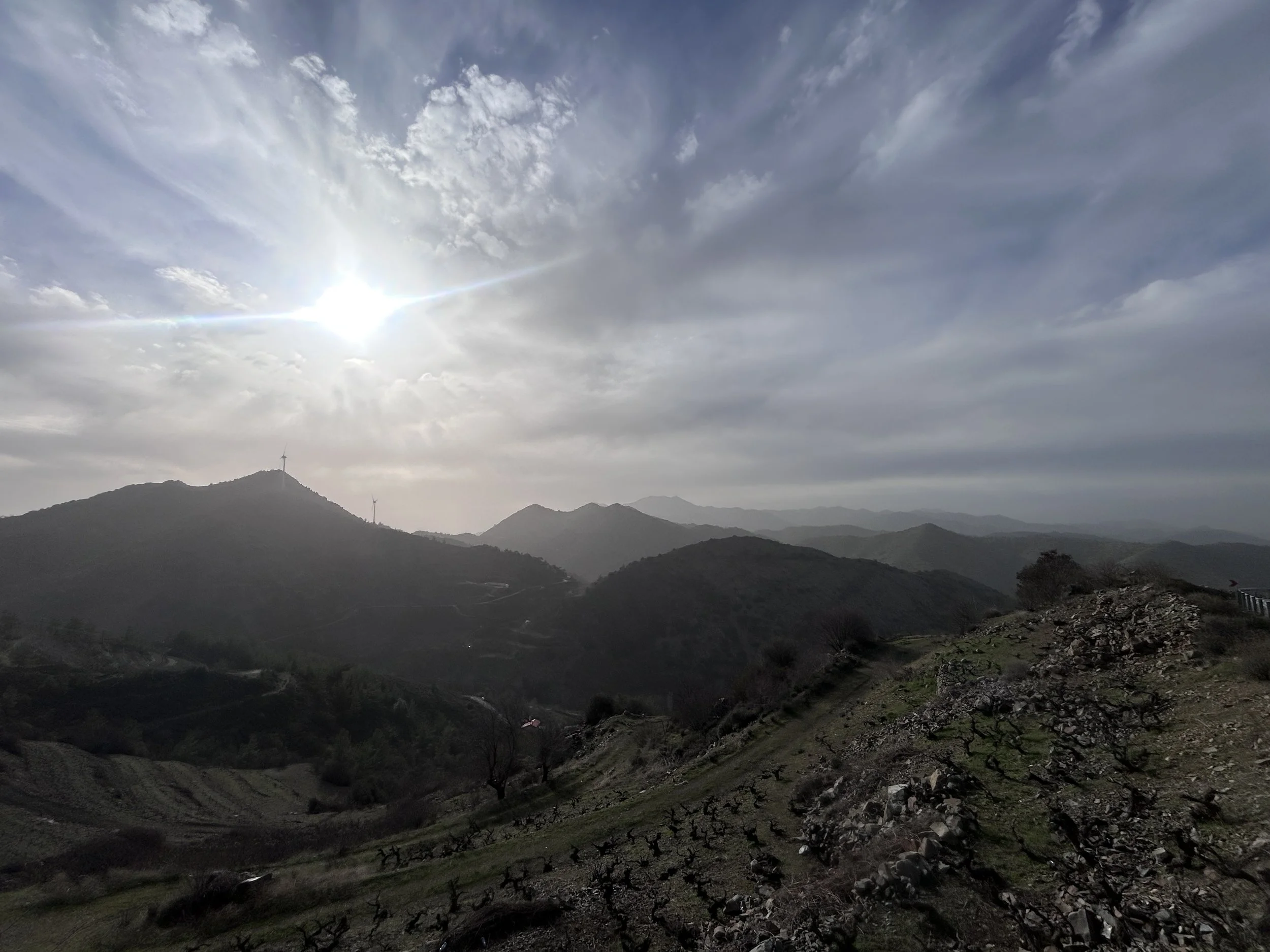 Mountain landscape with cloudy sky and bright sun, distant wind turbines, and terraced hills with sparse vegetation.