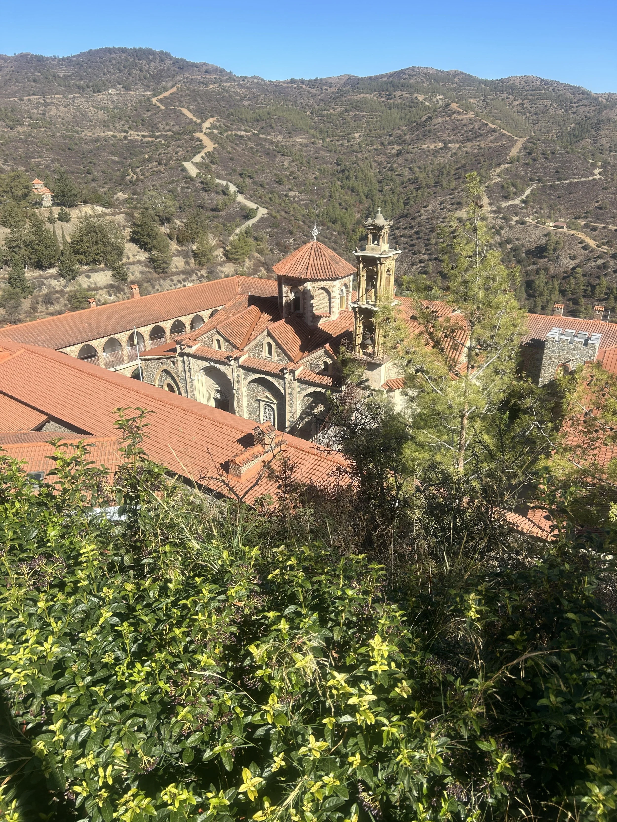 A church with red-tiled roofs, surrounded by greenery and mountains in the background under a clear blue sky.