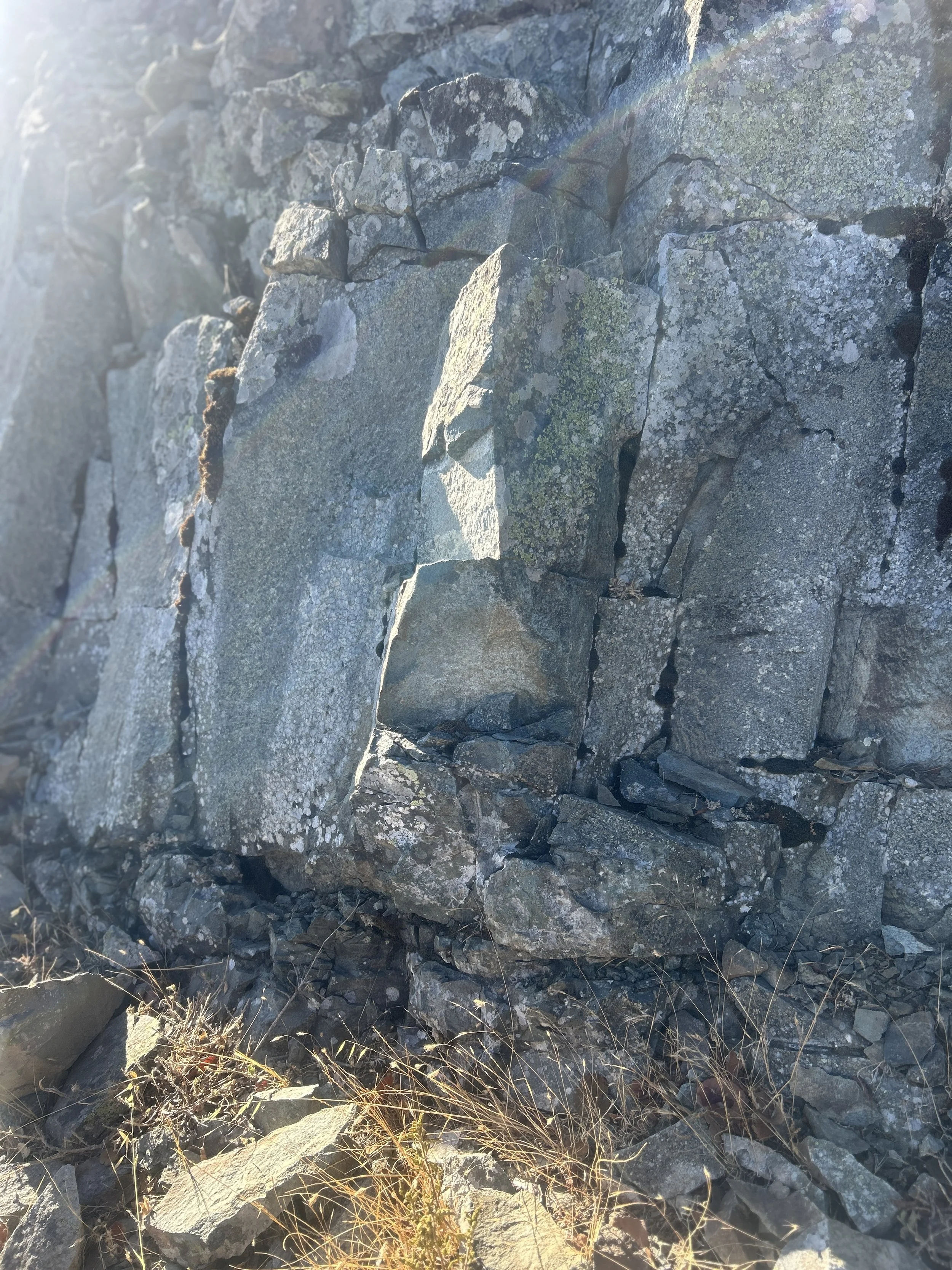 Close-up of a rock formation with gray and moss-covered stones, some with lichen, in bright sunlight with glare and a faint rainbow or rainbow-like light effect.