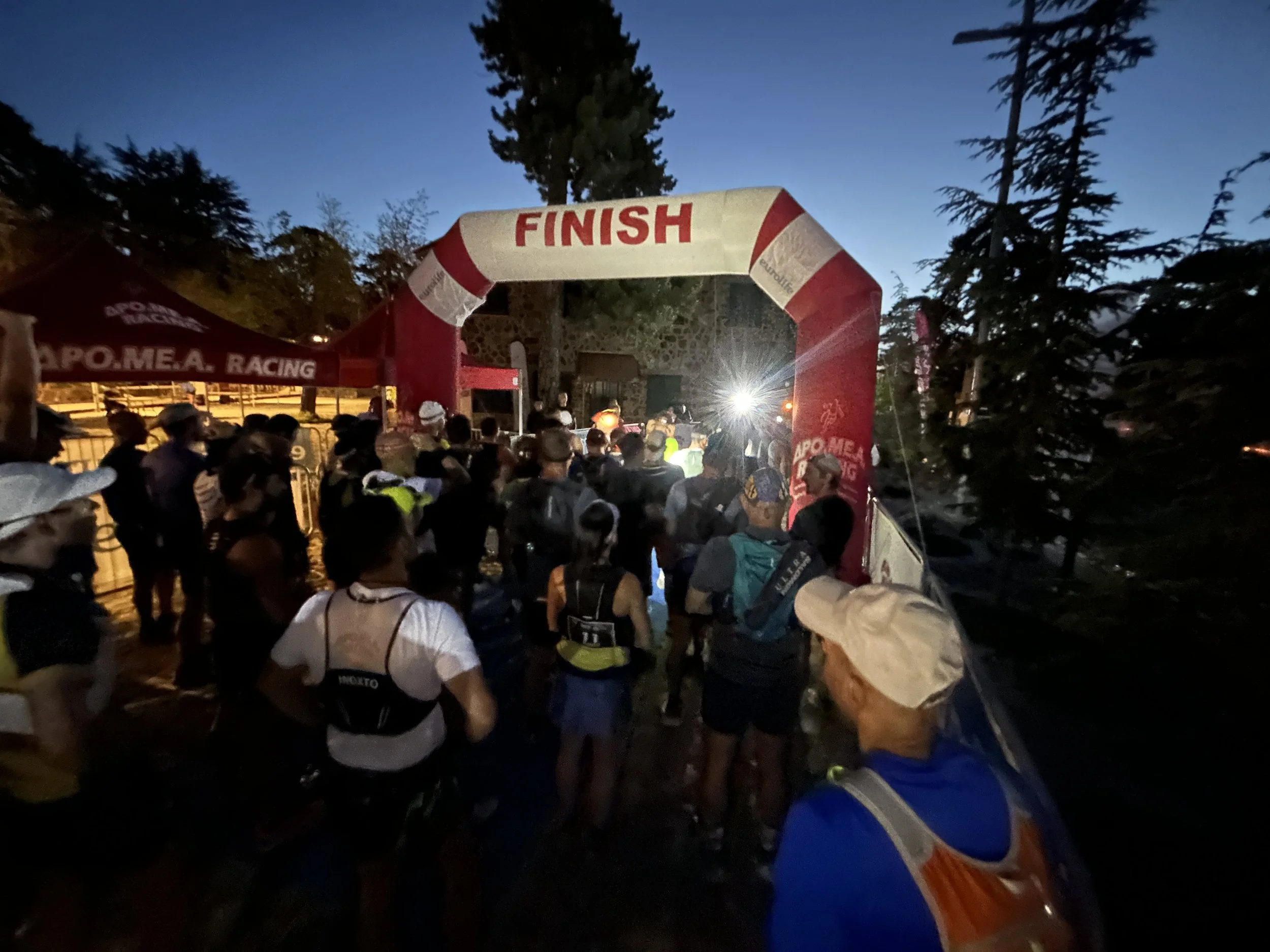 A large group of runners gathered under an arch that says 'FINISH' at a nighttime outdoor race event, with trees and tents in the background.
