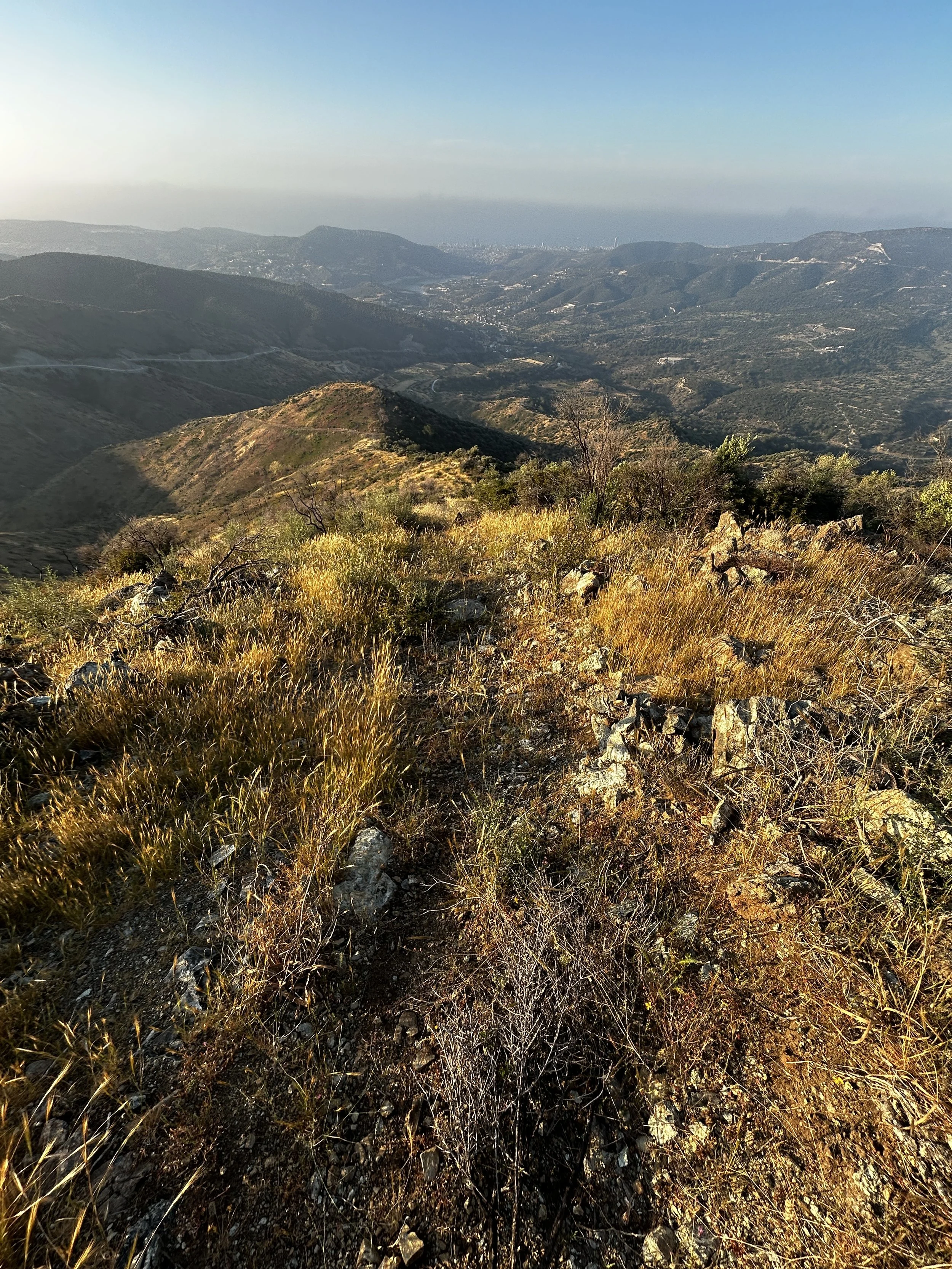 Mountain landscape with dry grass and rocks in foreground, rolling hills and valleys in middle, distant city skyline on horizon, clear sky with some haze.