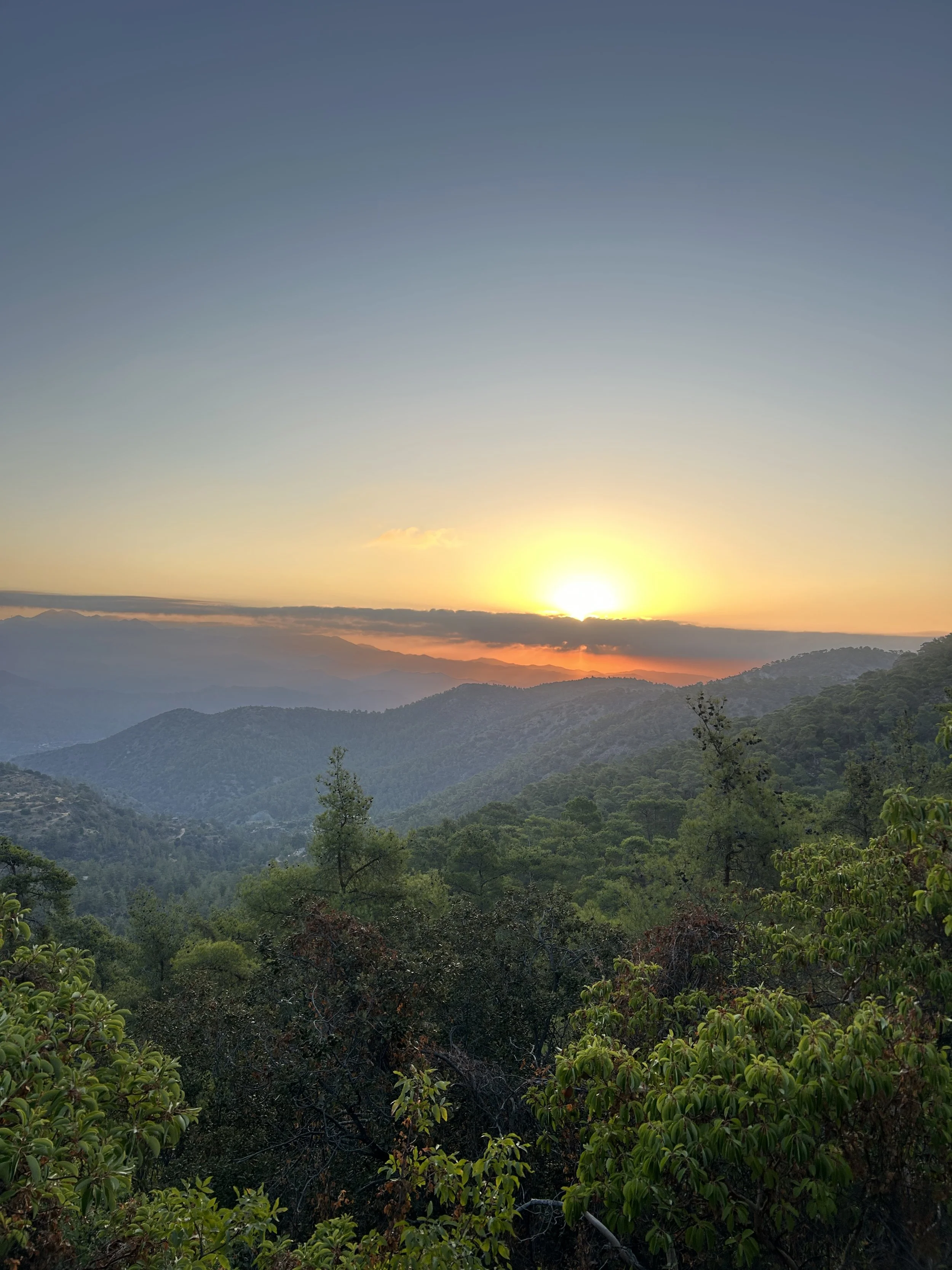 Sunset over a mountain range with layers of hills covered in green trees.
