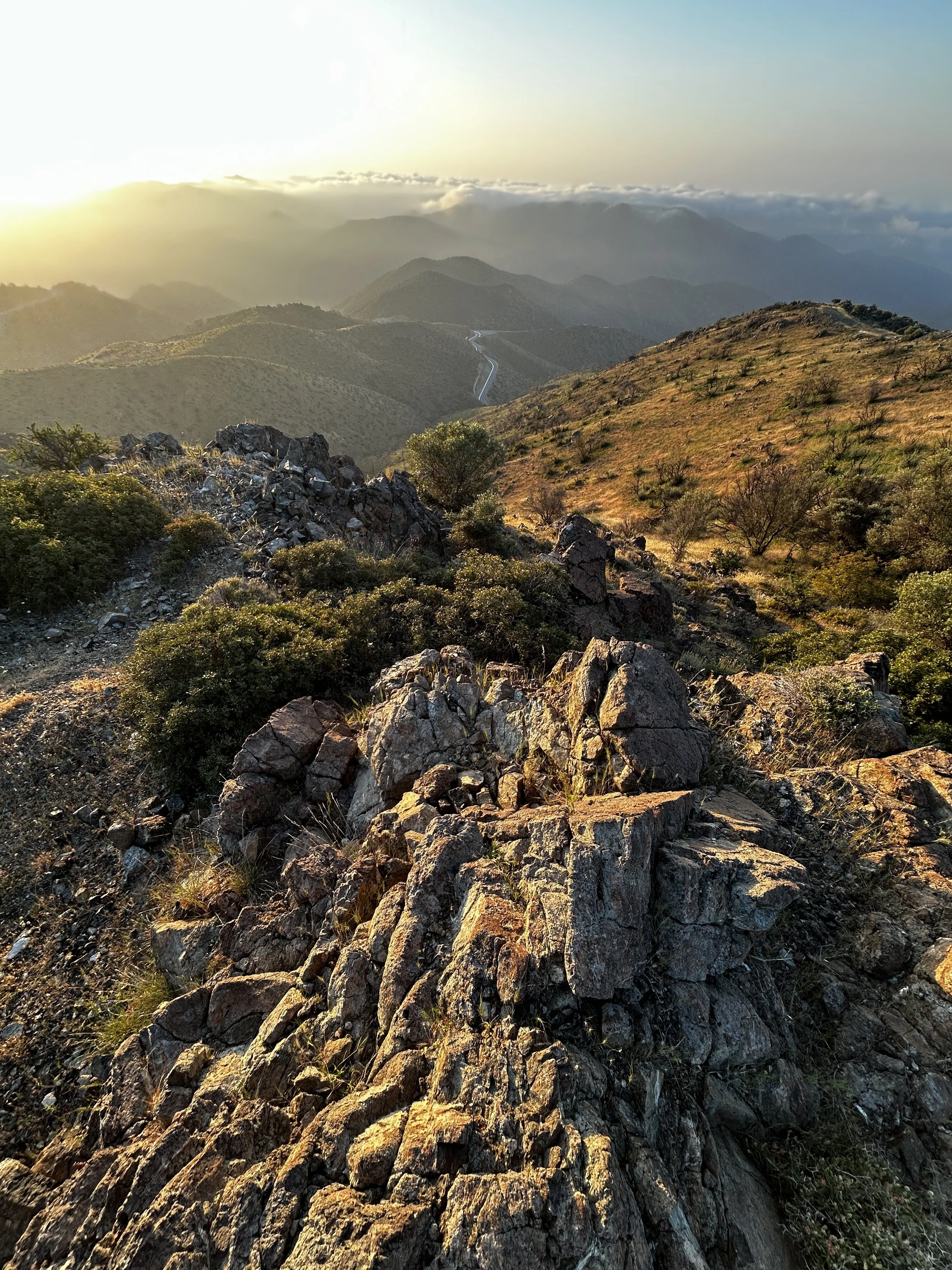 Sunset over a mountainous landscape with rocks and sparse shrubbery in the foreground.