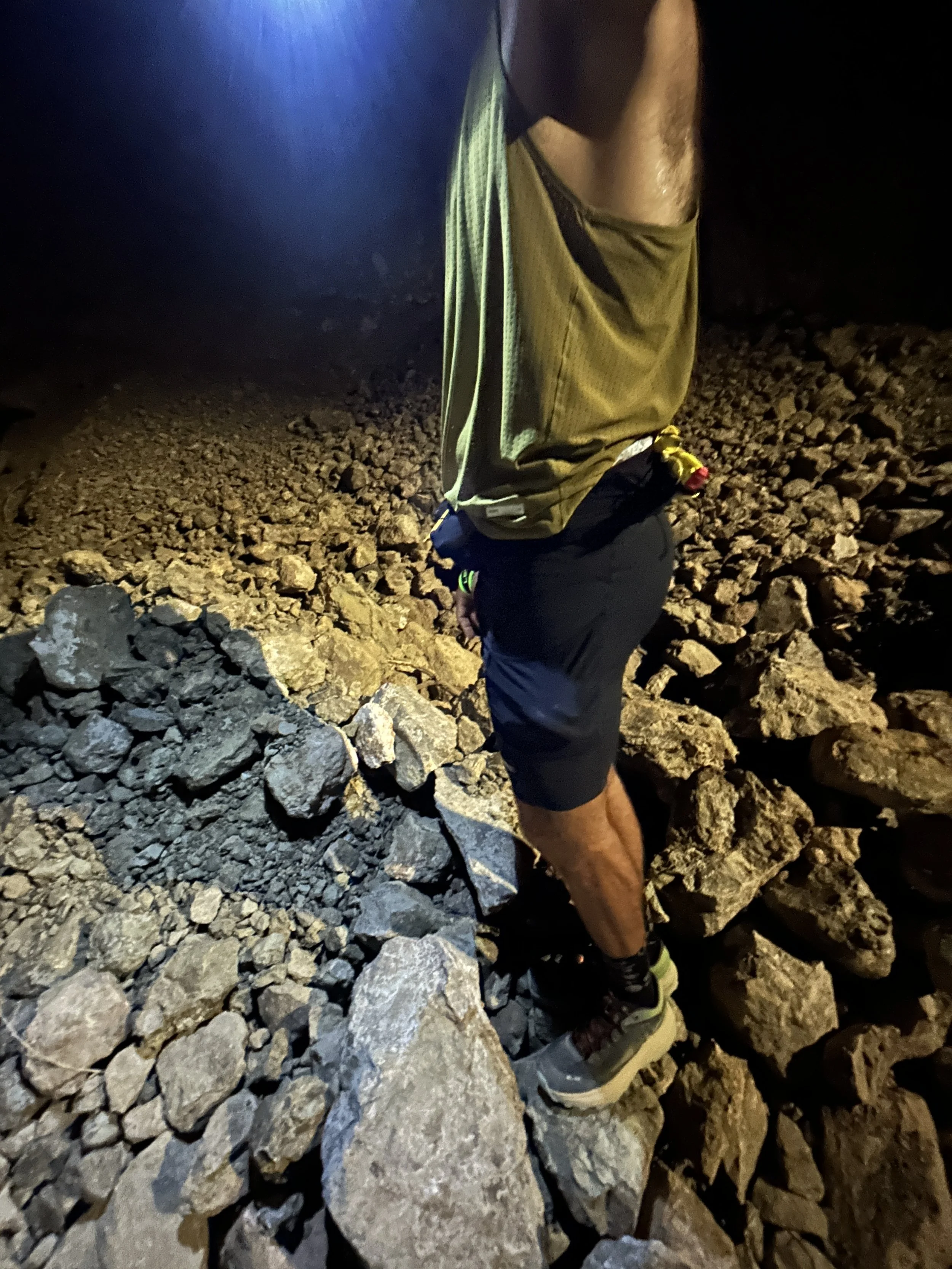 A person wearing trail running shoes, black shorts, and a tan shirt, standing on a rocky surface at night with a headlamp shining on the ground.