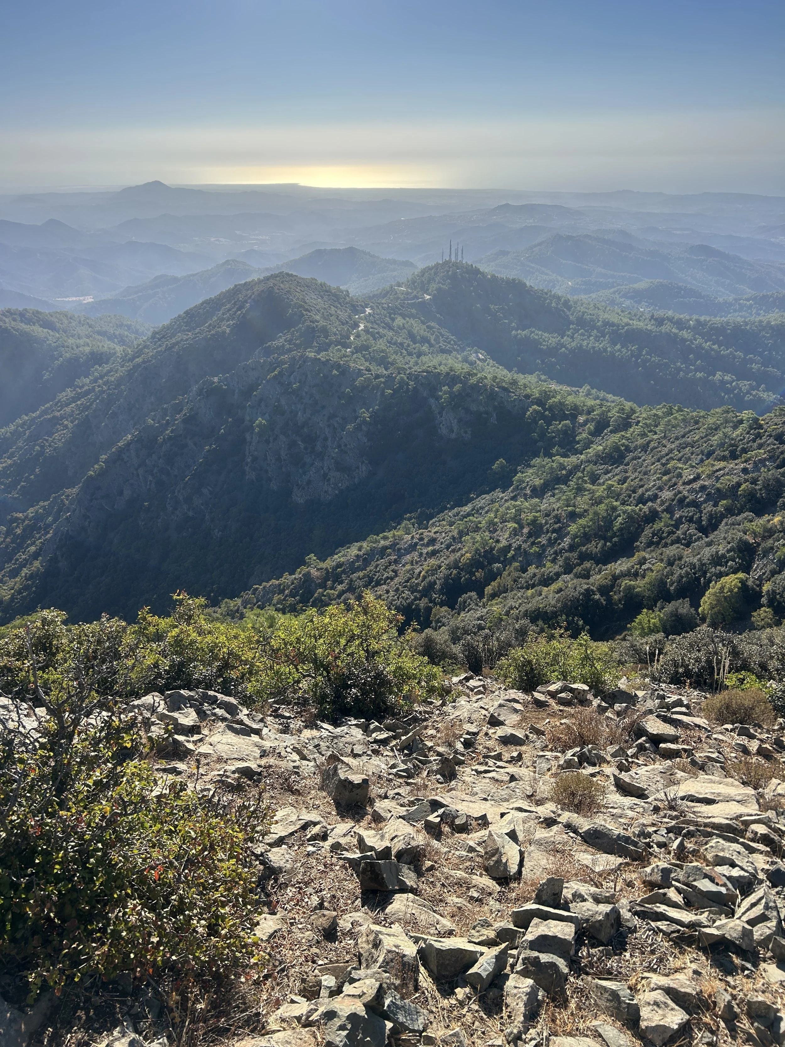 Mountain landscape with rocky terrain in the foreground, lush green hills and ridges in the middle ground, and a hazy horizon with the sun setting or rising in the background.