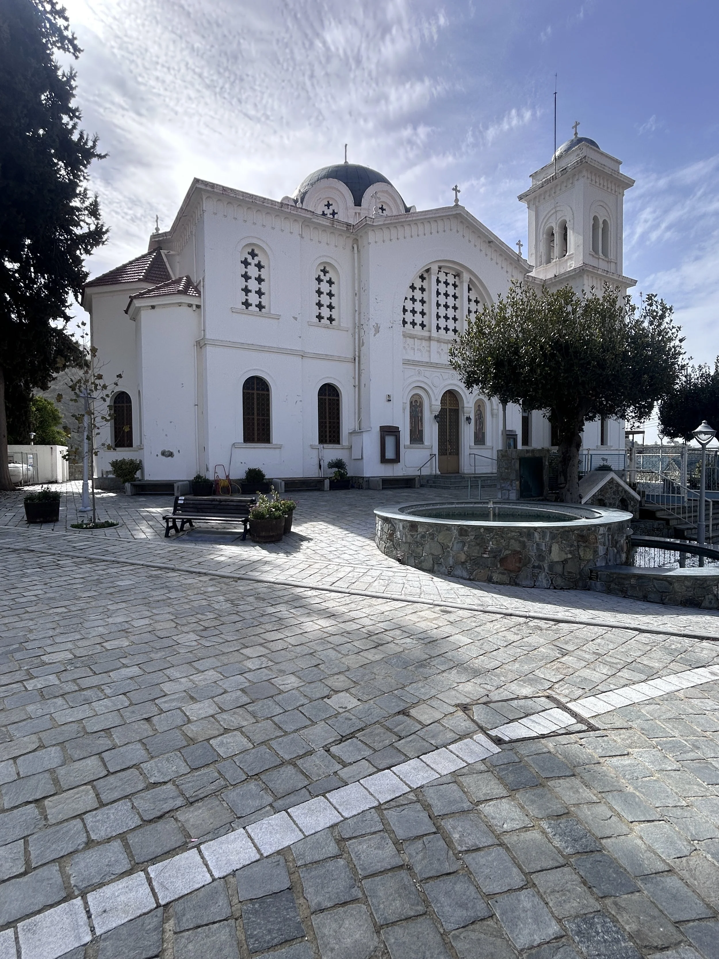 A white church with rounded windows, a dome, and a bell tower, surrounded by a paved courtyard with benches, potted plants, and a small fountain, under a partly cloudy sky.