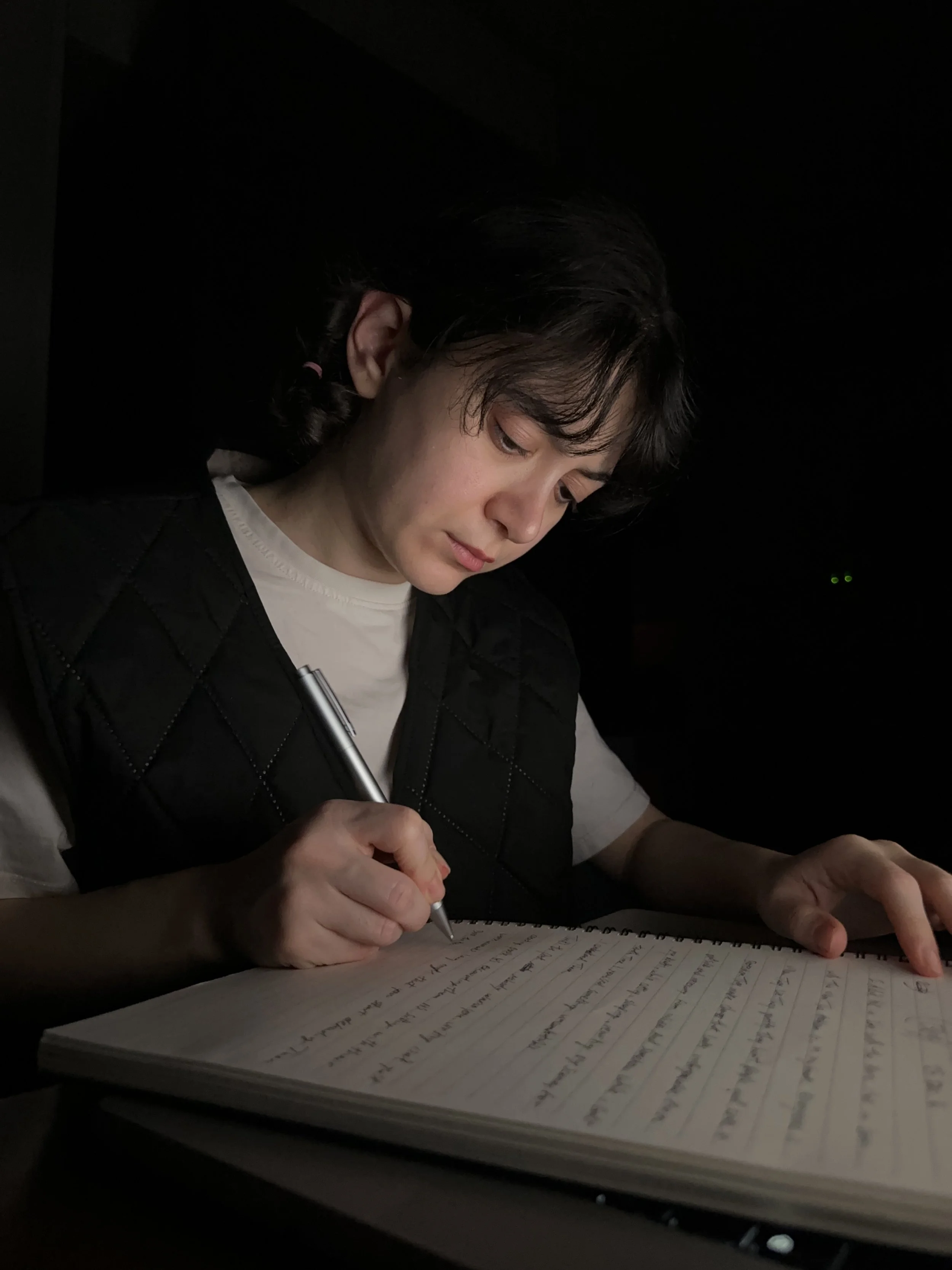 Young woman with short dark hair writing in a notebook with a pen, in a dimly lit environment.