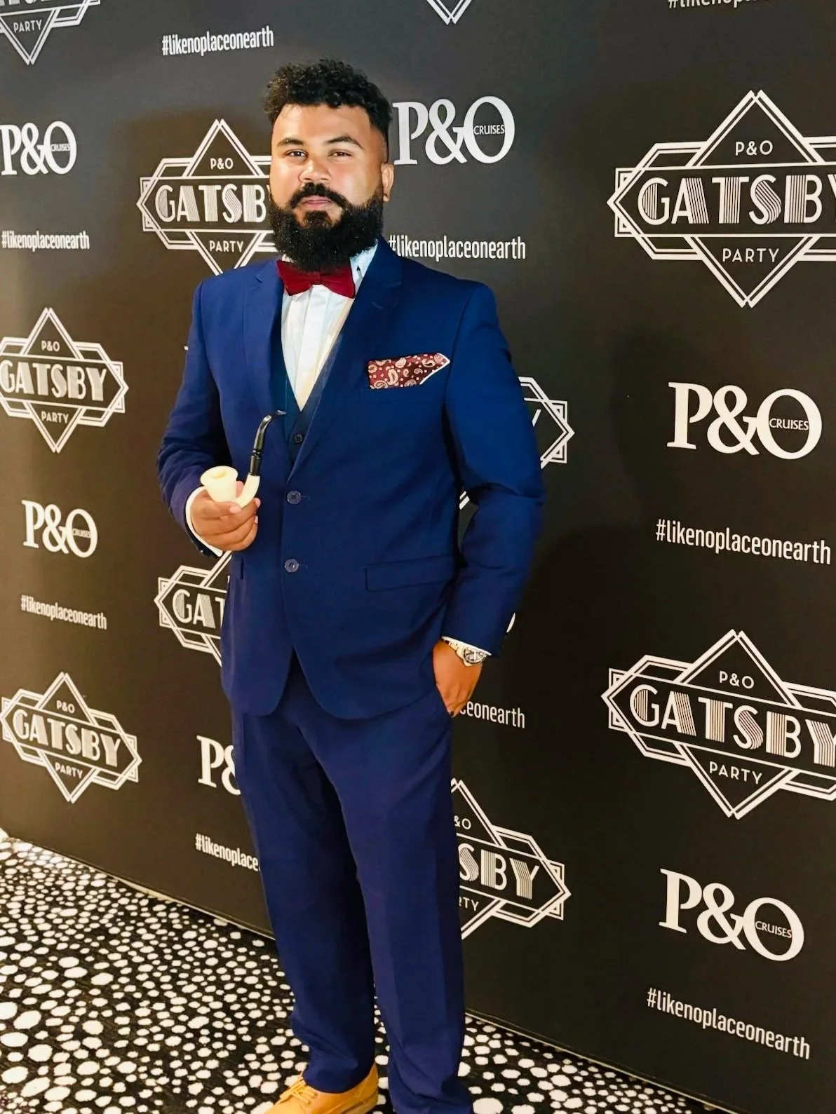 Man in a blue suit holding a pipe, standing on a black and white dotted carpet, at a Gatsby-themed party backdrop.