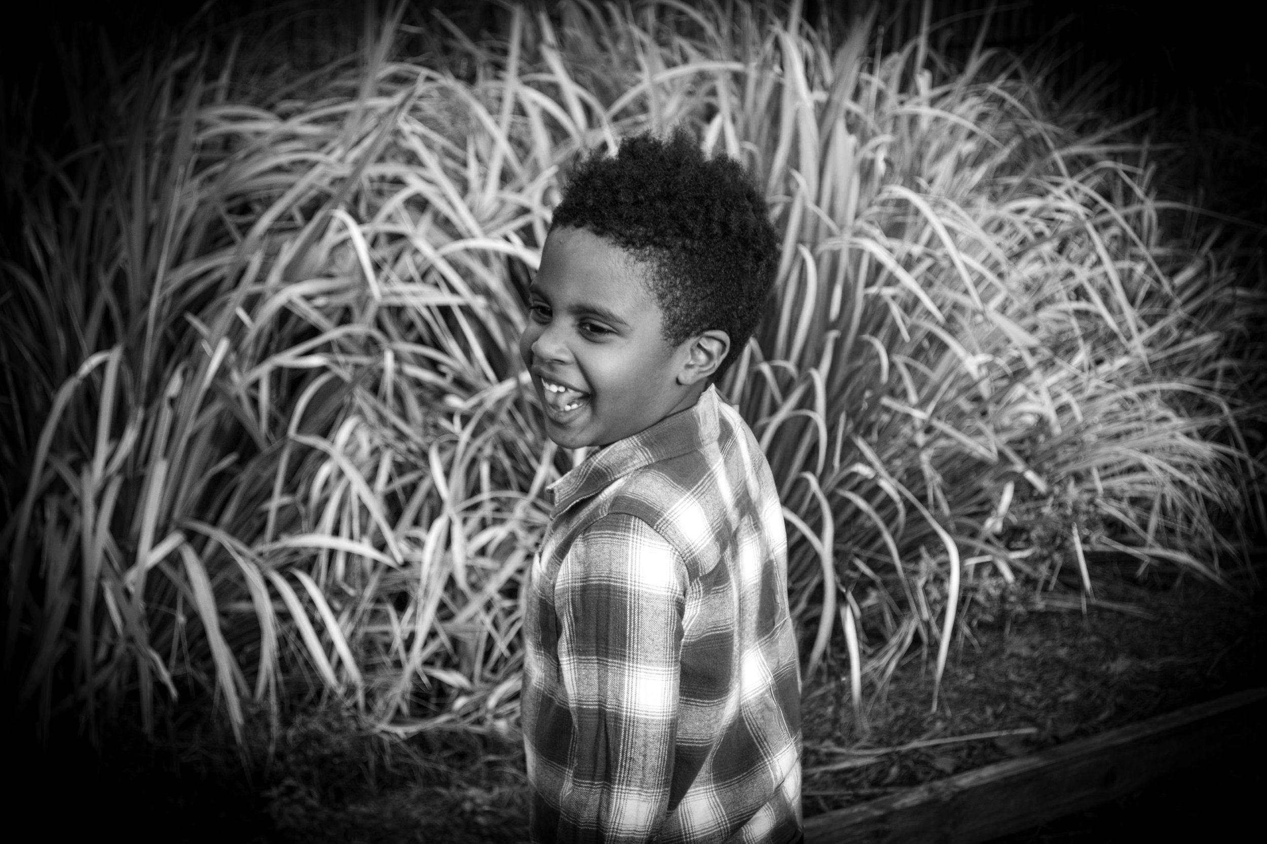 A young boy with curly hair wearing a checkered shirt smiling and walking outdoors past tall grass or plants.