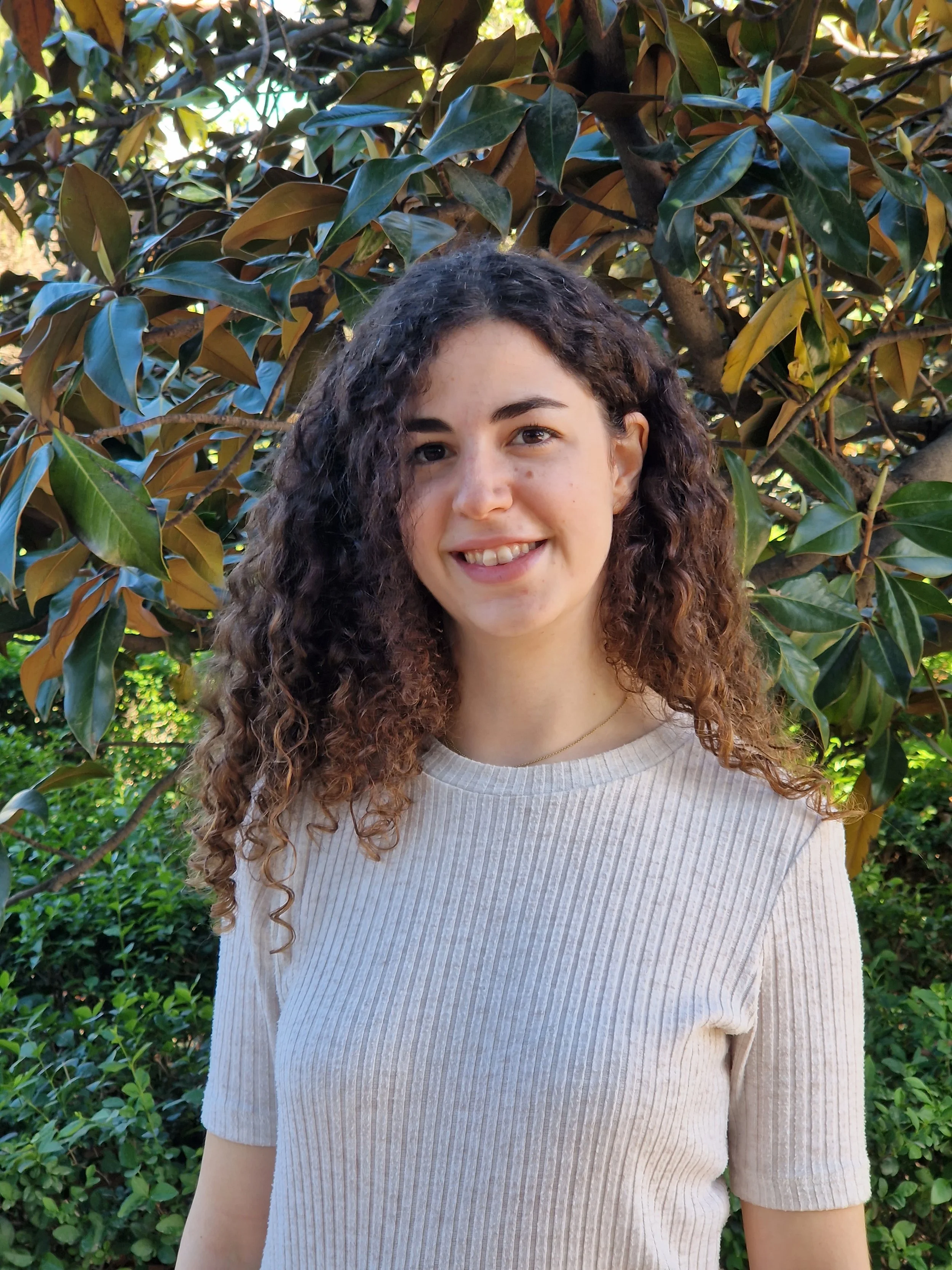 Fotografía de la psicóloga. Mujer joven con cabello rizado sonriendo, vestida con suéter de tono crema, en un entorno natural con plantas y árboles verdes y hojas grandes.