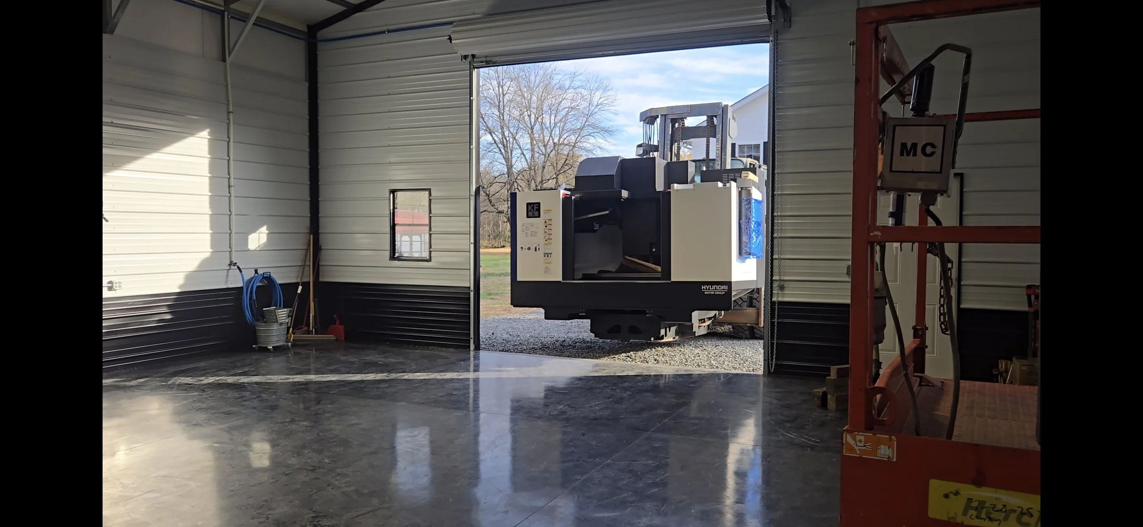 View of the inside of a spacious metal garage with an open door. Outside, a hydraulic lift is visible through the open doorway, parked on a gravel surface with trees and a building in the background. Inside the garage, there are cleaning supplies and equipment on the left, and a red lift on the right.