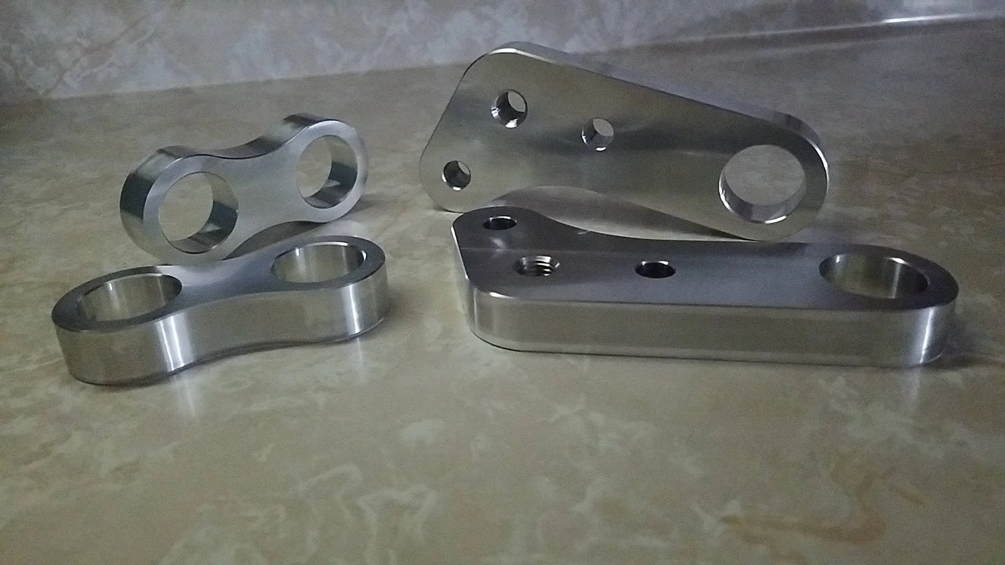 Four machined metal parts with holes, placed on a beige surface.