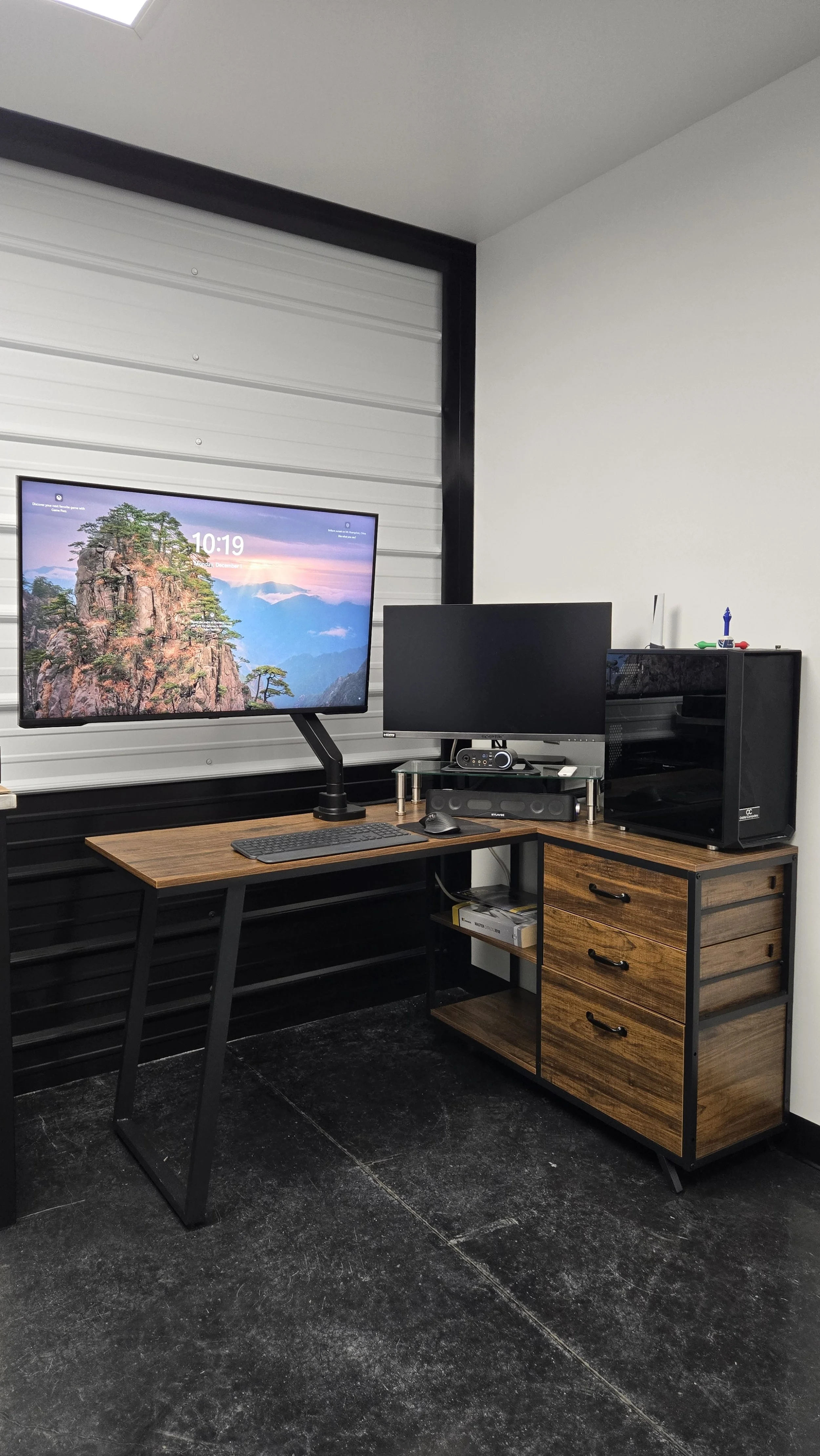 A home office desk setup with two monitors, a keyboard, mouse, a computer tower, and shelves on a black desk, with a white and black wall and a textured floor.