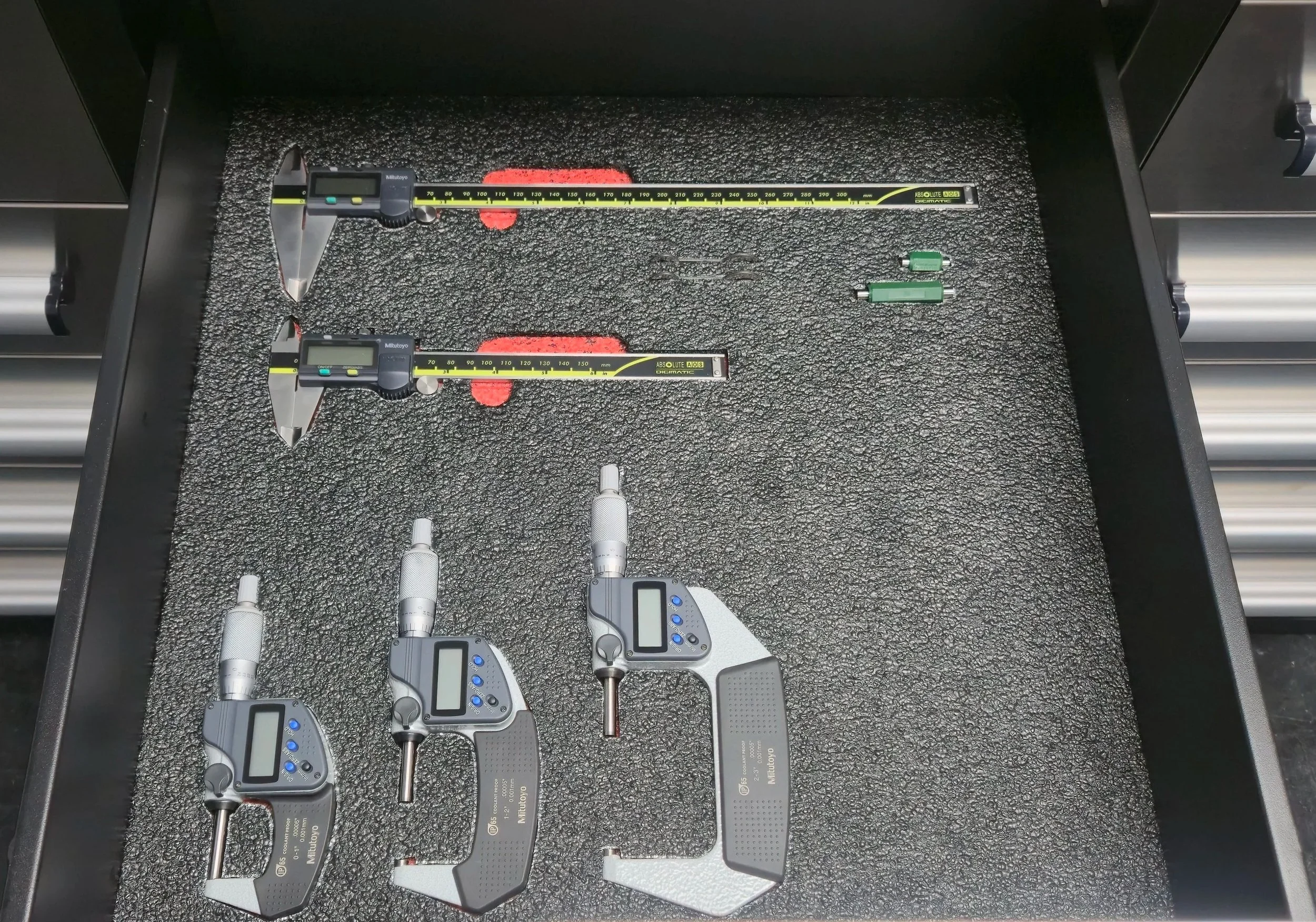 Three digital micrometers, two linear measurement tools, and two green electrical connectors on a textured black surface inside a toolbox drawer.
