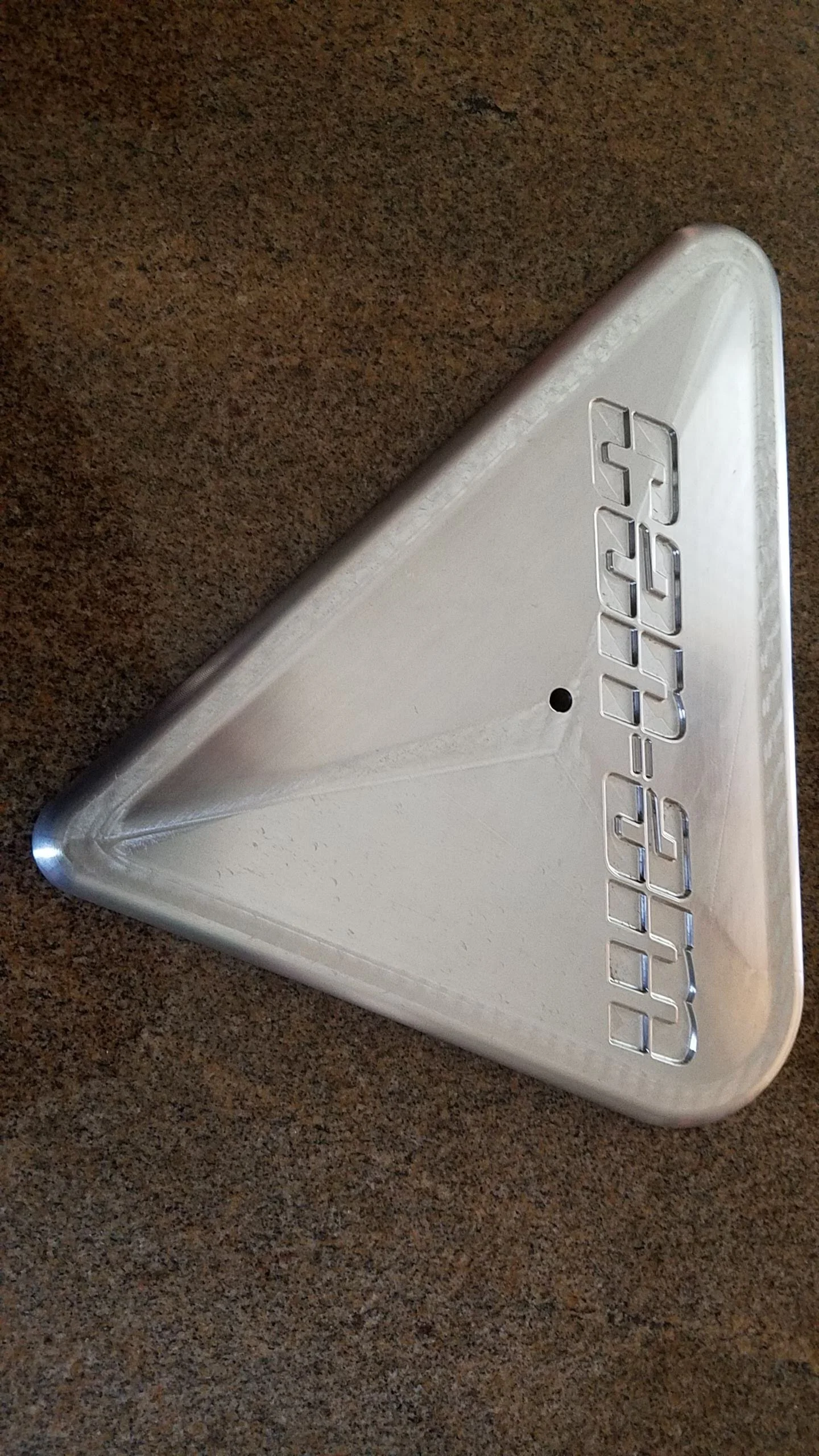 Close-up of a silver, triangular-shaped metal object with the word 'GATORADE' embossed in reverse on it, placed on a brown textured surface.