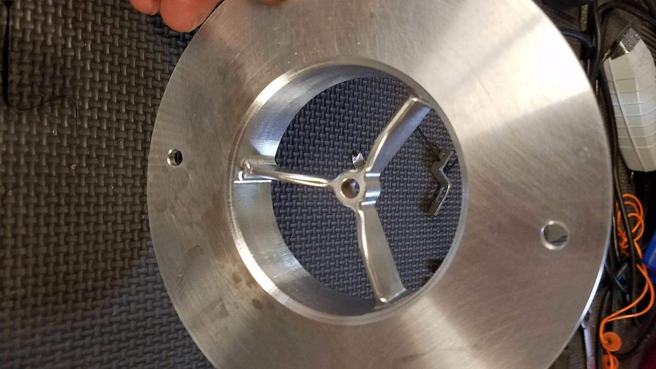 A circular metal component with a three-arm spider and a central hole, resting on a black textured surface. There are small holes on the perimeter of the component, and some wires are visible in the background.