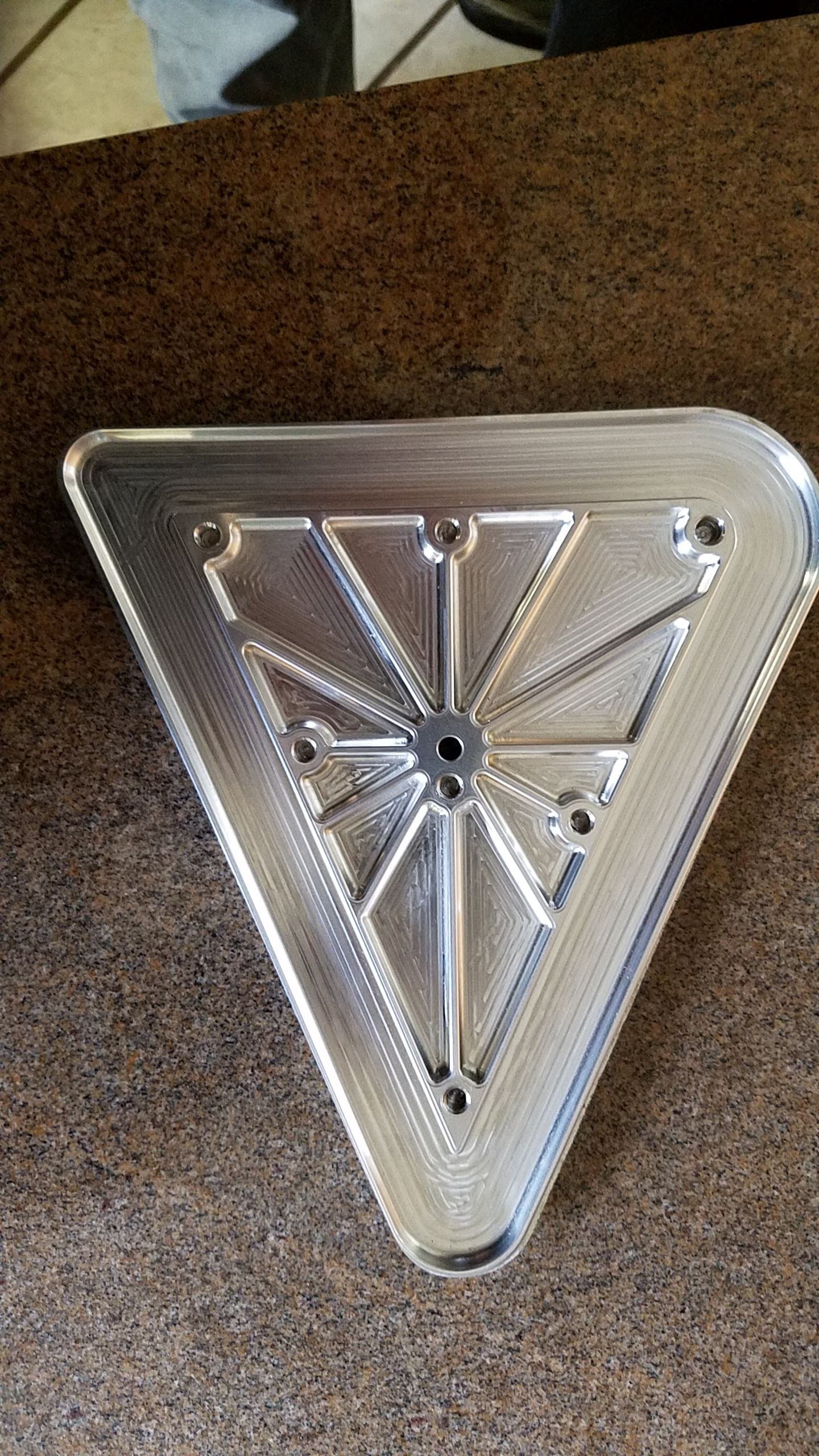 A triangular metallic object with intricate grooves and a central hole, resting on a brown textured surface.