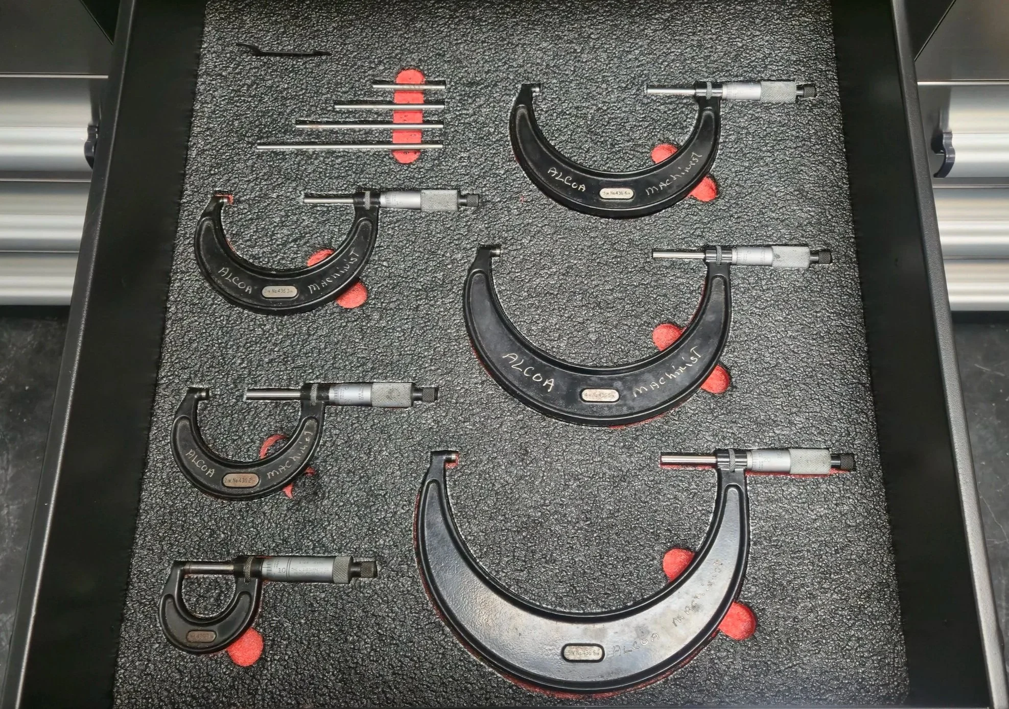 Set of six black dial calipers with silver measuring arms, arranged on a foam tray with red foam pads, with some calipers labeled 'ALcoa' and 'Machinist'.