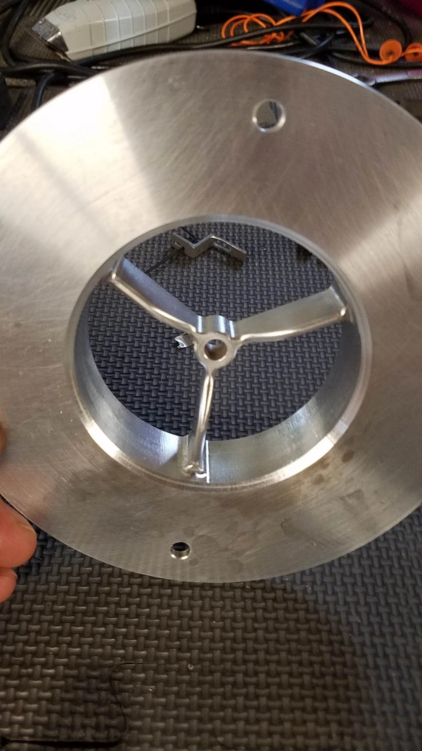 A close-up of a metallic mechanical device with a circular opening, featuring a clock hand and internal components, placed on a textured dark surface.