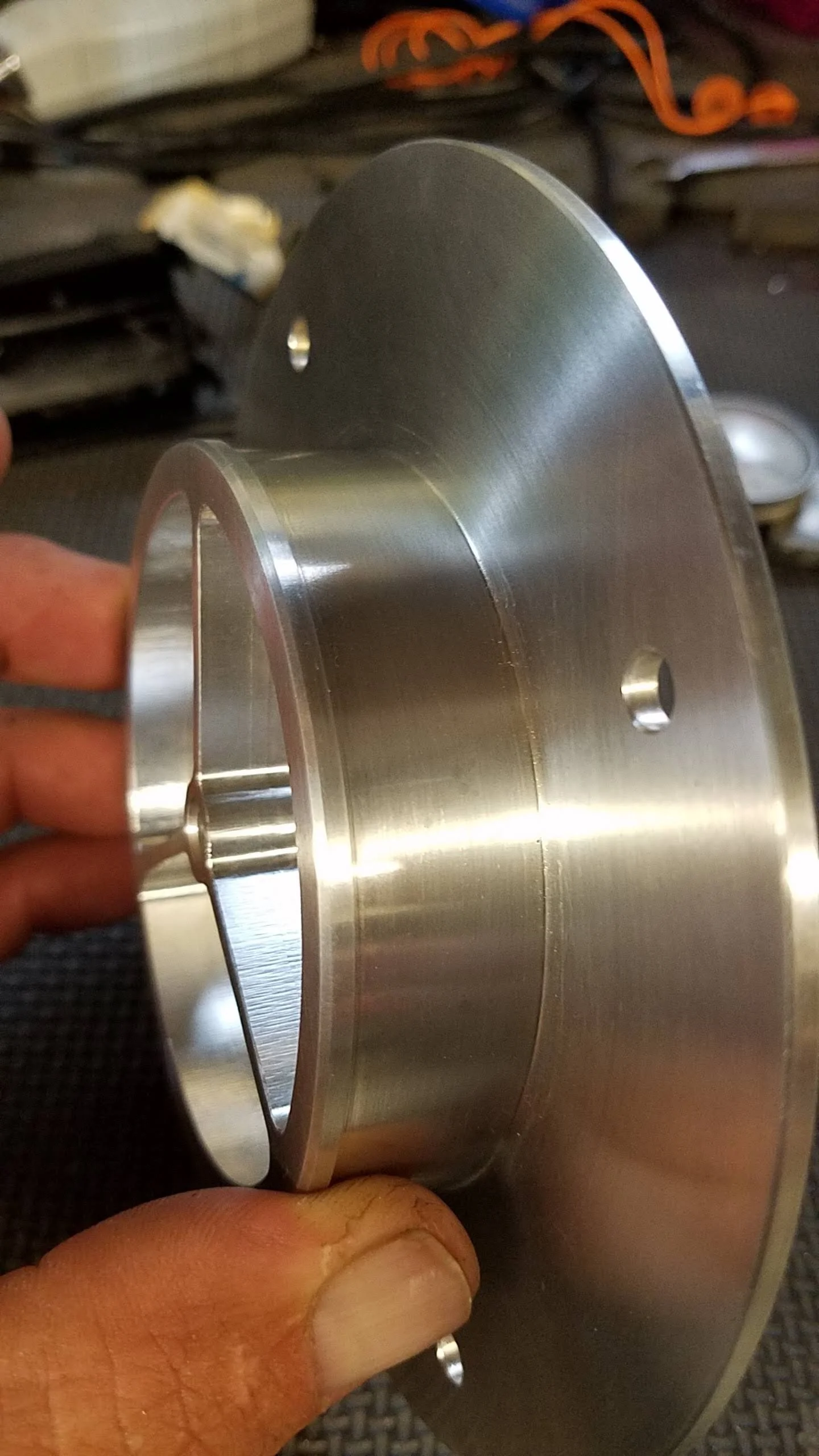 A person holding a machined metal wheel or pulley with a smooth, shiny surface and holes around its edge, likely made of aluminum or stainless steel.