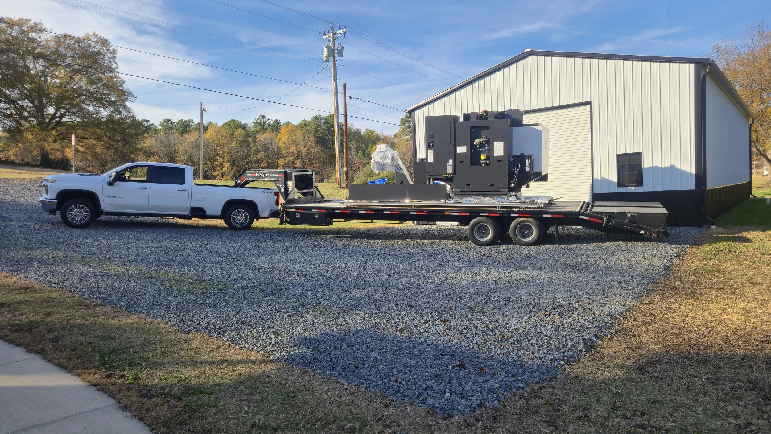 White pickup truck towing a flatbed trailer loaded with a large black industrial machine parked outside a metal-sided building, with a tree and power lines in the background.