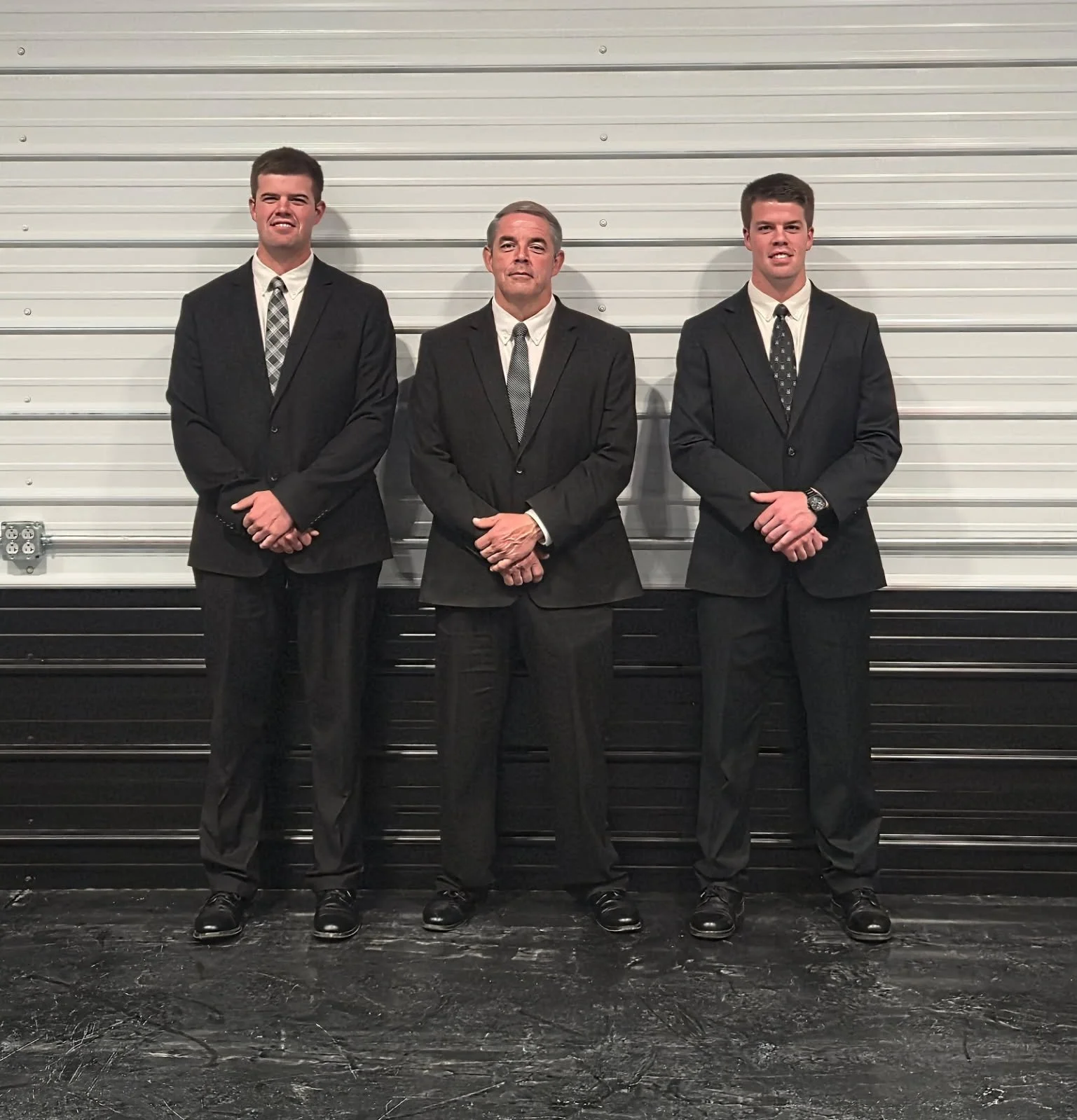 Three men in black suits standing against a white garage door, with serious and smiling expressions.