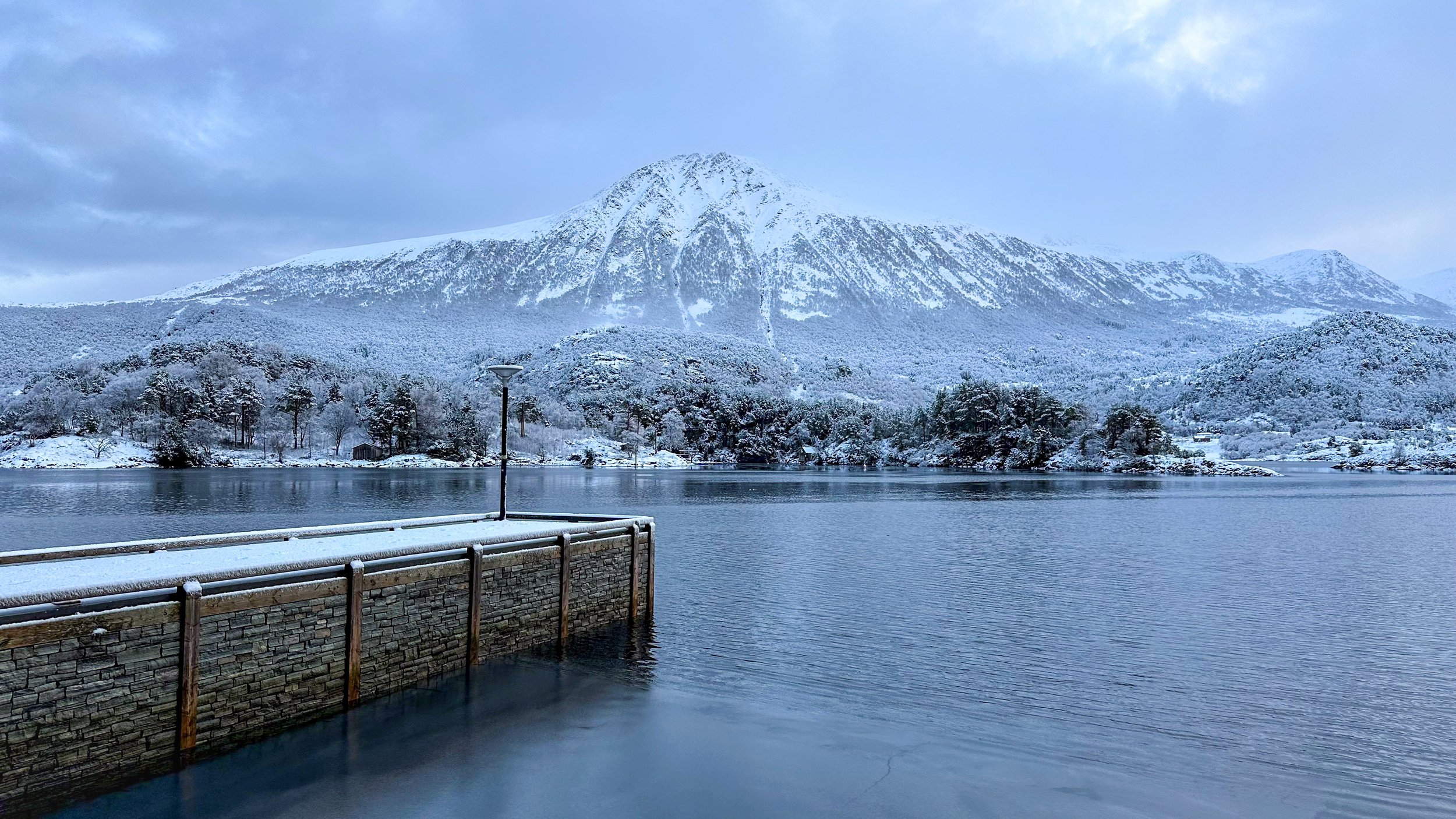 Snow-covered mountain in the background with a partly cloudy sky, trees along the shoreline, a lake in the foreground, and a small dock with a lamppost covered in snow.