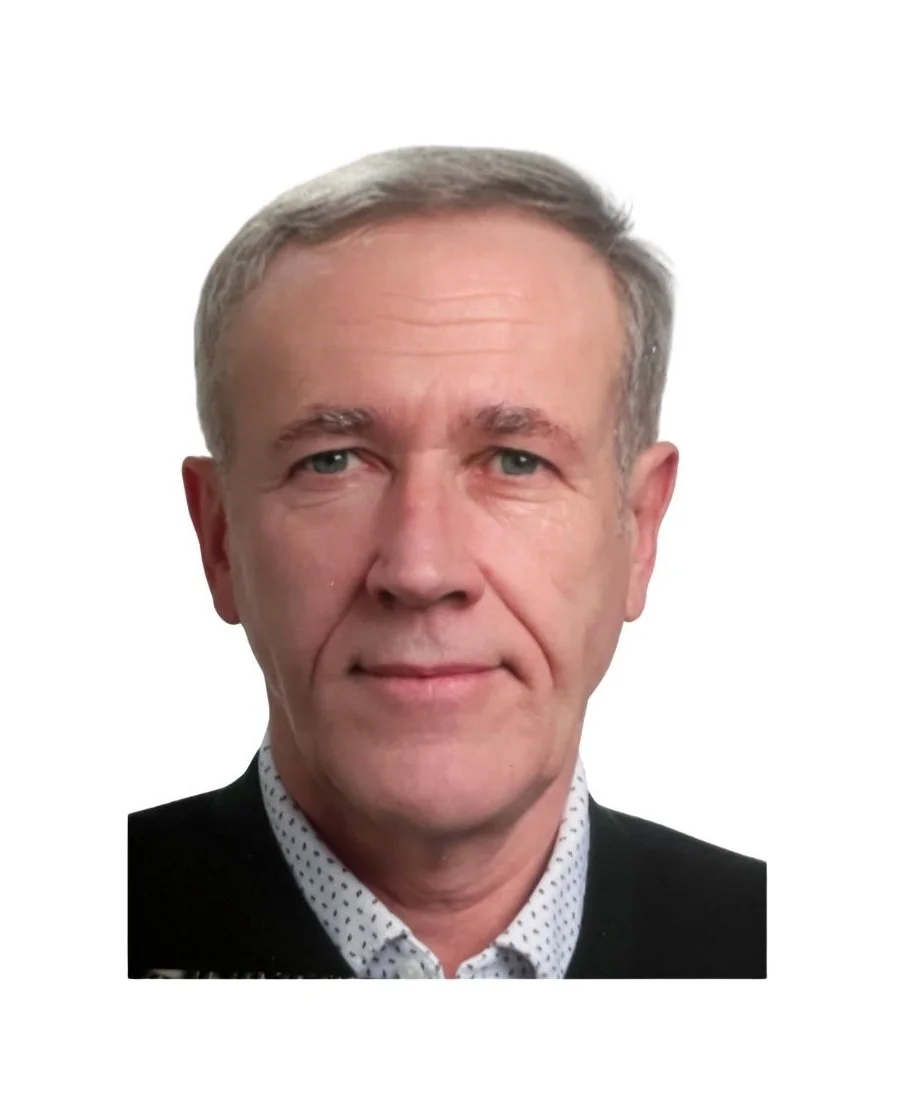 Porträt eines Mannes mit grauem Haar und blauen Augen, trägt ein schwarzes Jackett und ein weißes Hemd mit Punkten, vor weißem Hintergrund.