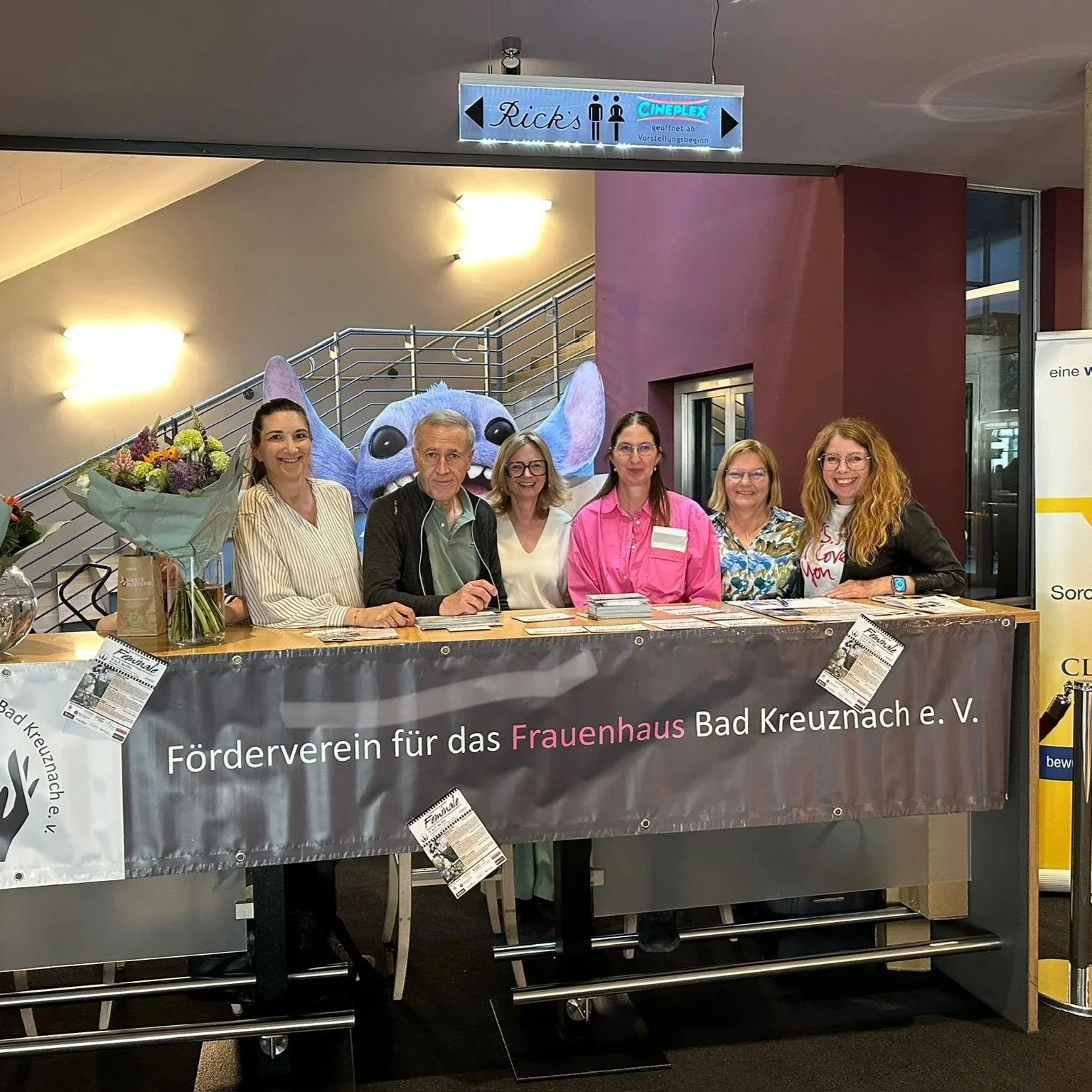 Sechs Frauen und ein Mann stehen hinter einem Tisch mit einem Banner, das den Förderverein für das Frauenhaus Bad Kreuznach e.V. unterstützt. Im Hintergrund ist eine Stickfigur von Stitch aus „Lilo & Stitch“ und eine Treppe sichtbar.
