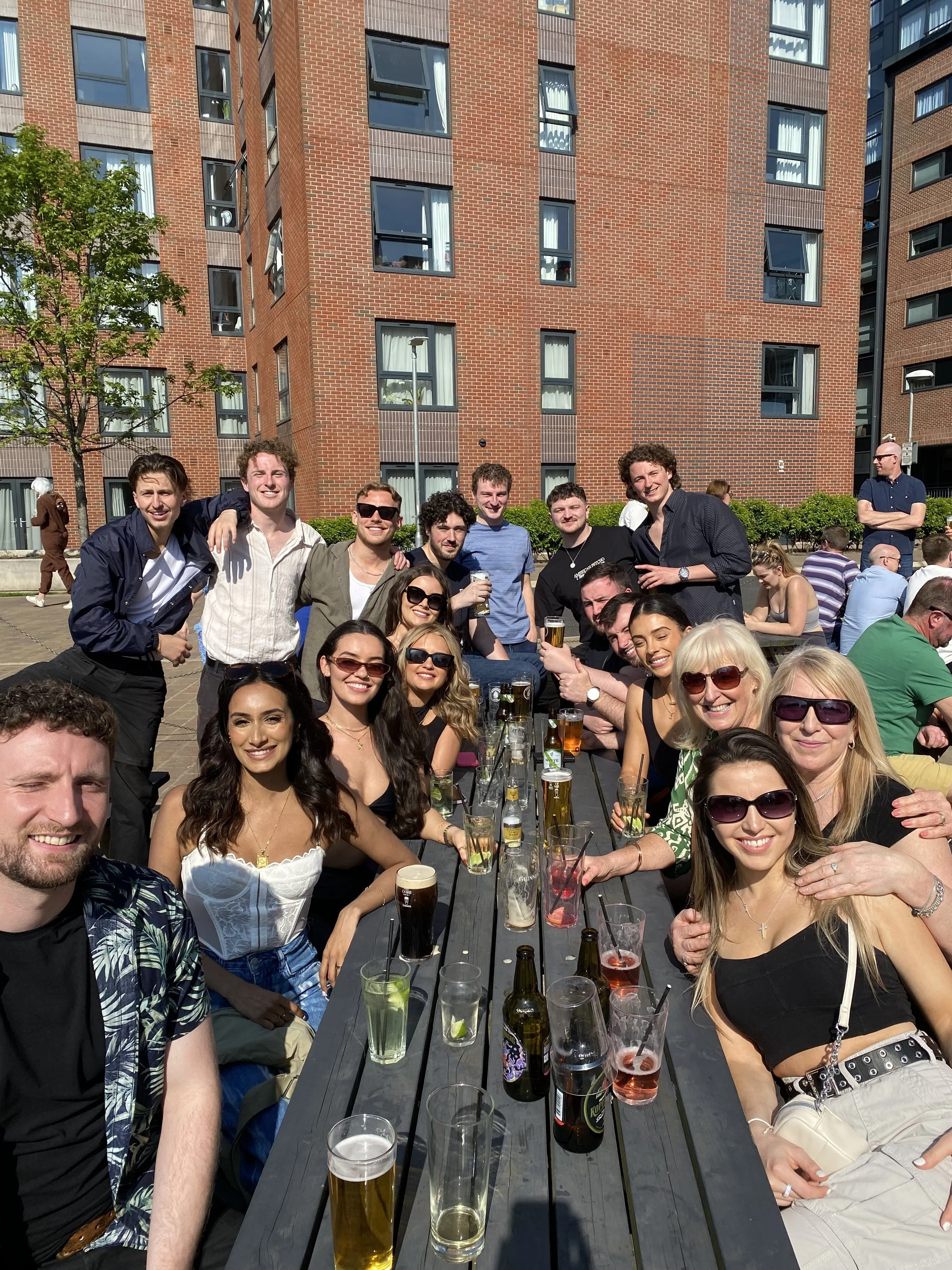 A group of smiling young adults gathered around a long outdoor table with drinks, enjoying a sunny day in an urban setting with brick buildings in the background.