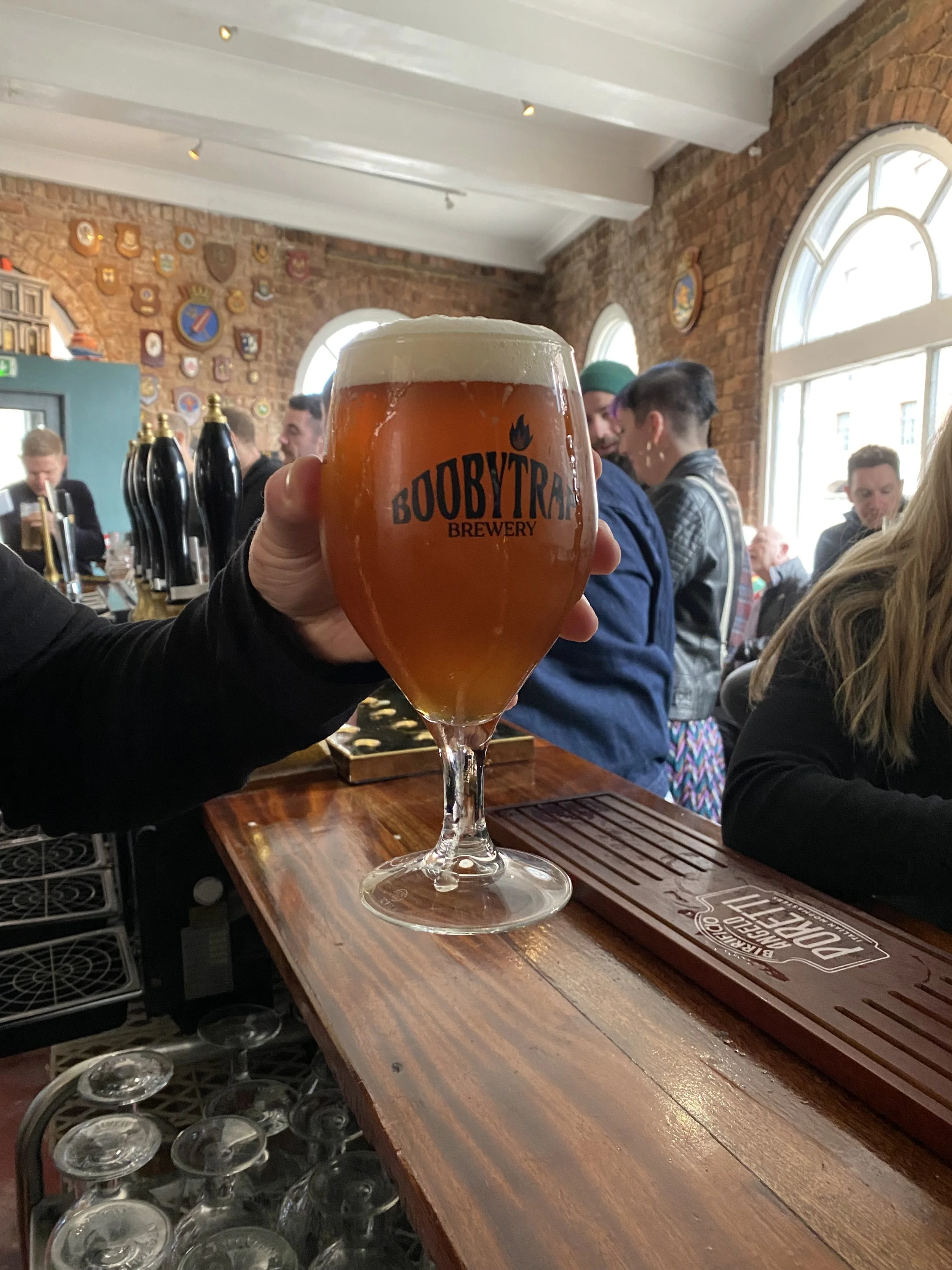 A person holding a glass of amber beer with a foamy head, branded with 'BOOBYTRAP BREWERY,' in a cozy brewery or pub with brick walls, large arched windows, and decorative shields on the wall. The bar has a wooden surface with bar equipment.