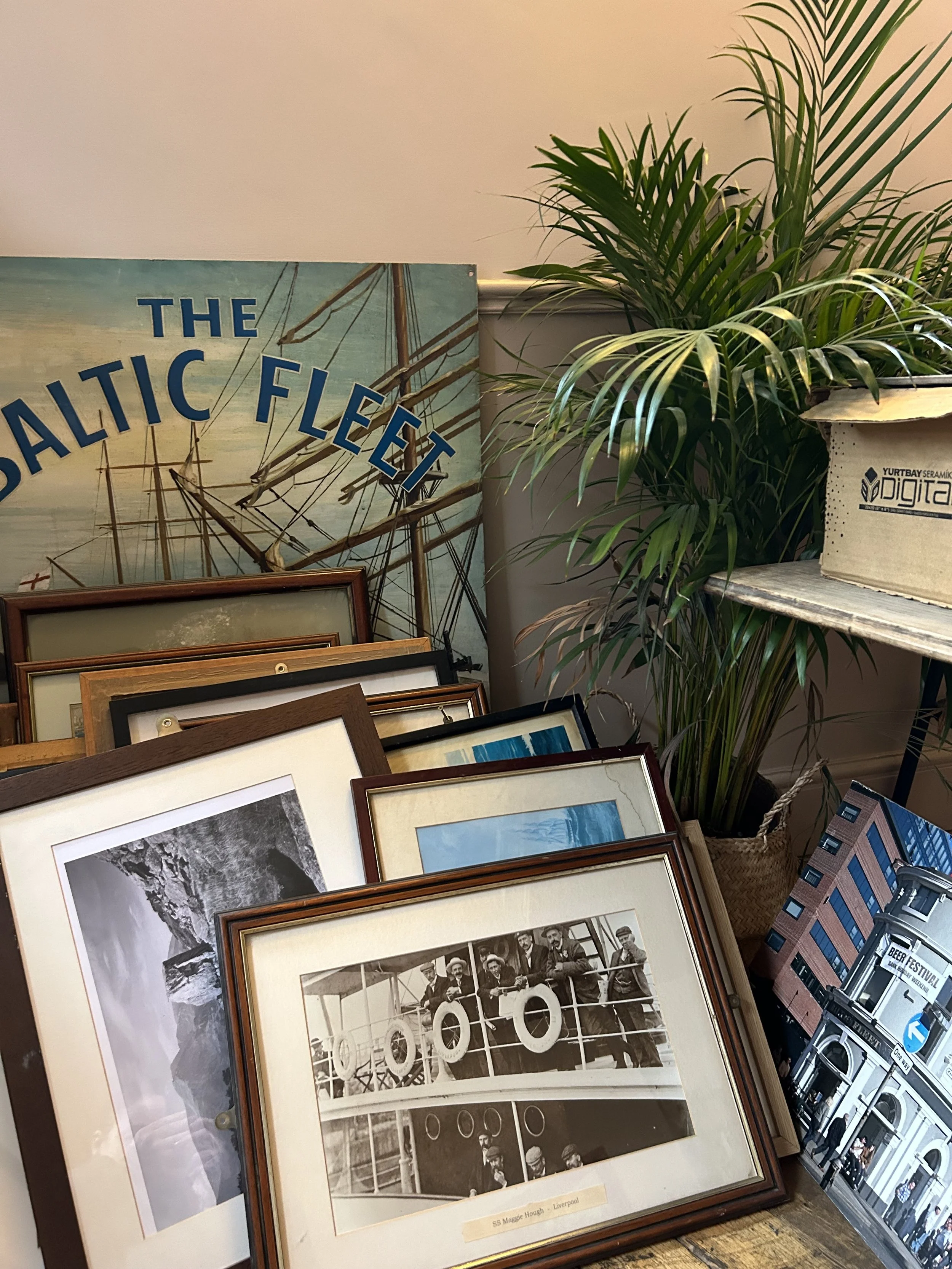 A collection of framed photographs and artwork, including a vintage photo of men on a ship, placed against a wall with a large sign that reads 'The Baltic Fleet.' A tall green plant is on the right, next to a cardboard box with the words 'Digital' an