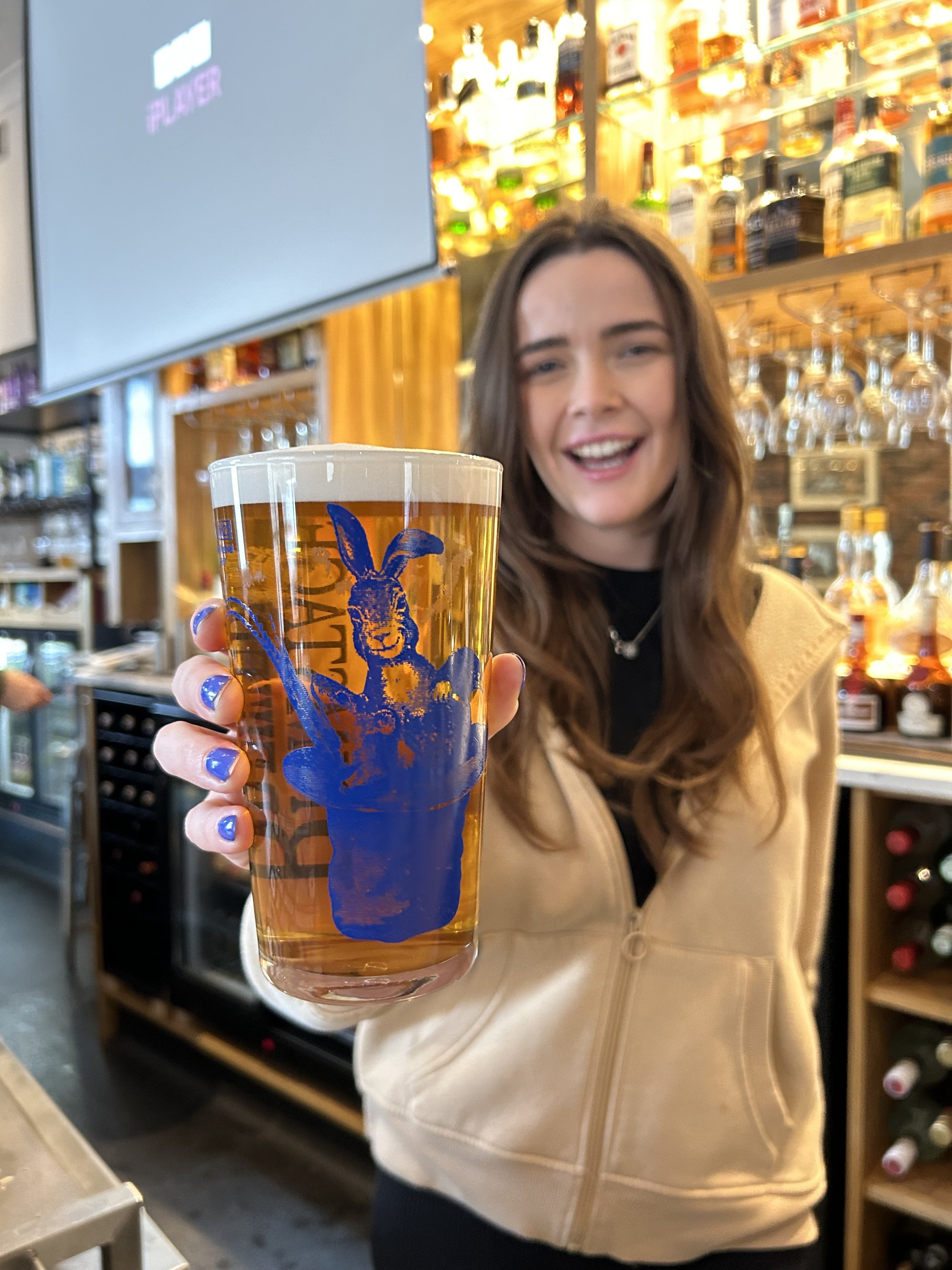 A young woman with long brown hair and a beige jacket holding a pint glass of beer with a blue logo of a rabbit in a bar or pub setting.
