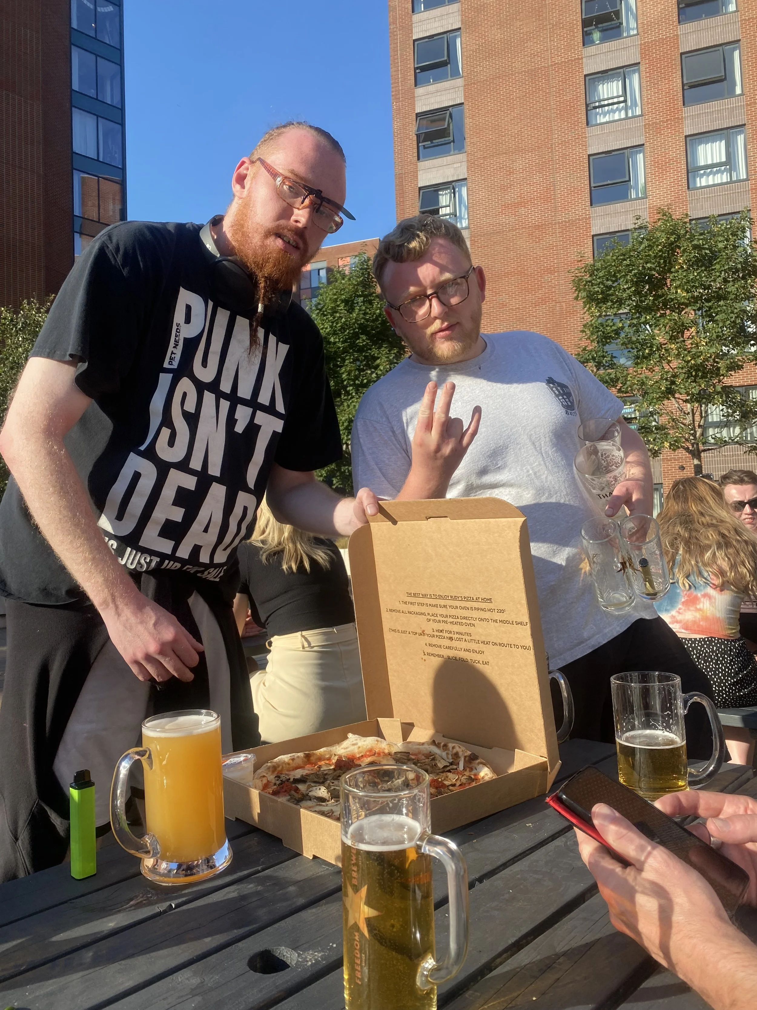 Two men at an outdoor gathering with pizza and drinks, one wearing a black t-shirt with bold white text and the other wearing glasses and a gray t-shirt, surrounded by people, with a city apartment building in the background.