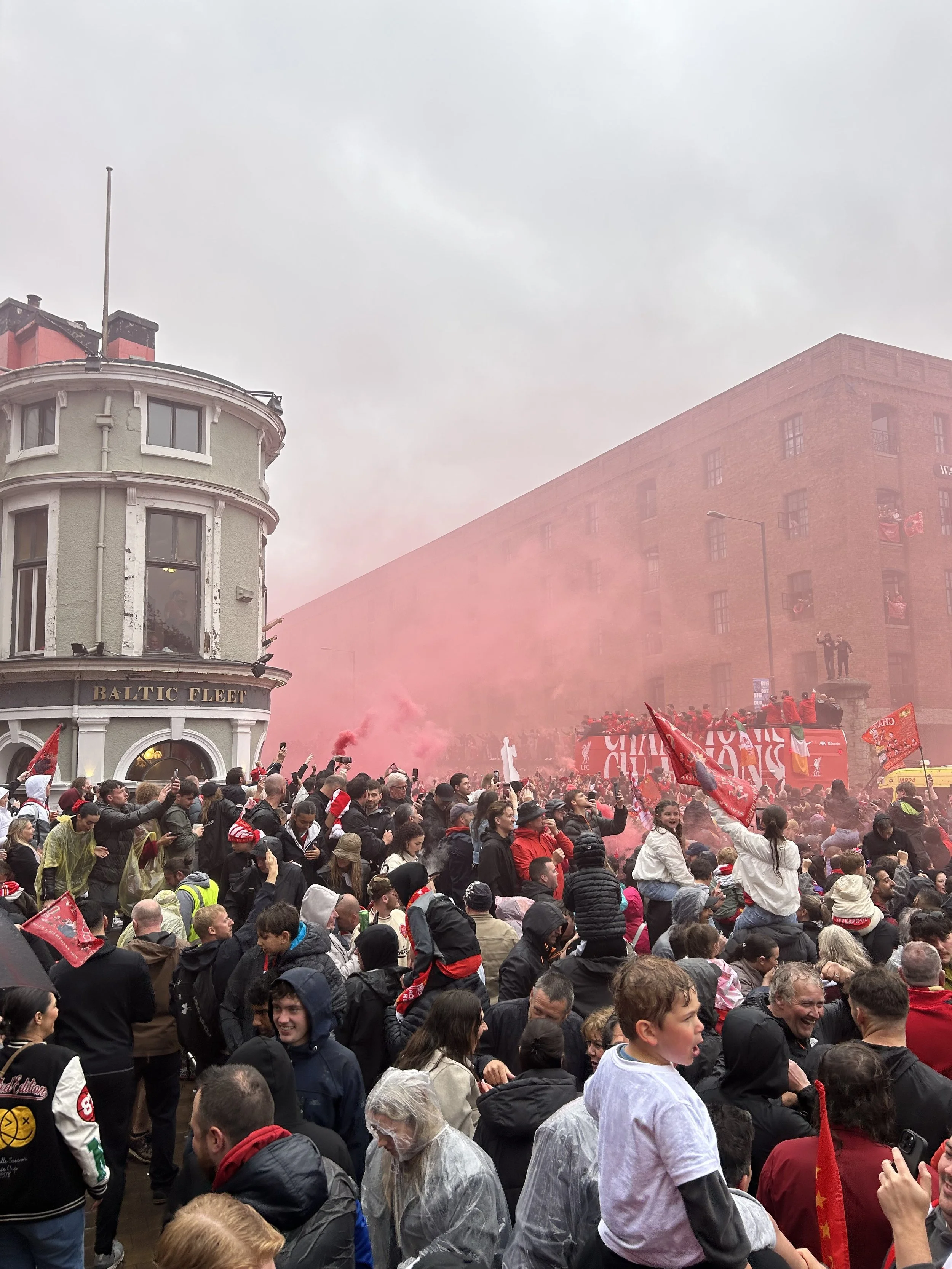 Crowd gathered on a city street during a celebration, with people waving flags, smoke, and banners, under overcast skies near buildings labeled 'Baltic Fleet'.