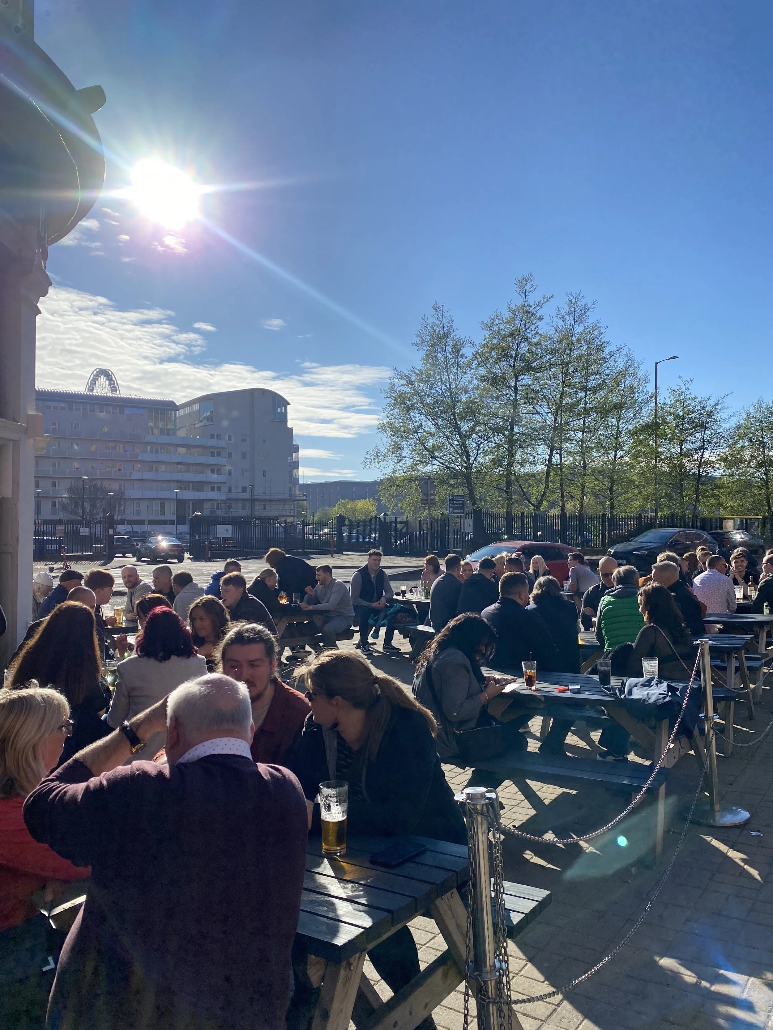 People dining outdoors at picnic tables on a sunny day with blue sky, some clouds, and trees in the background.