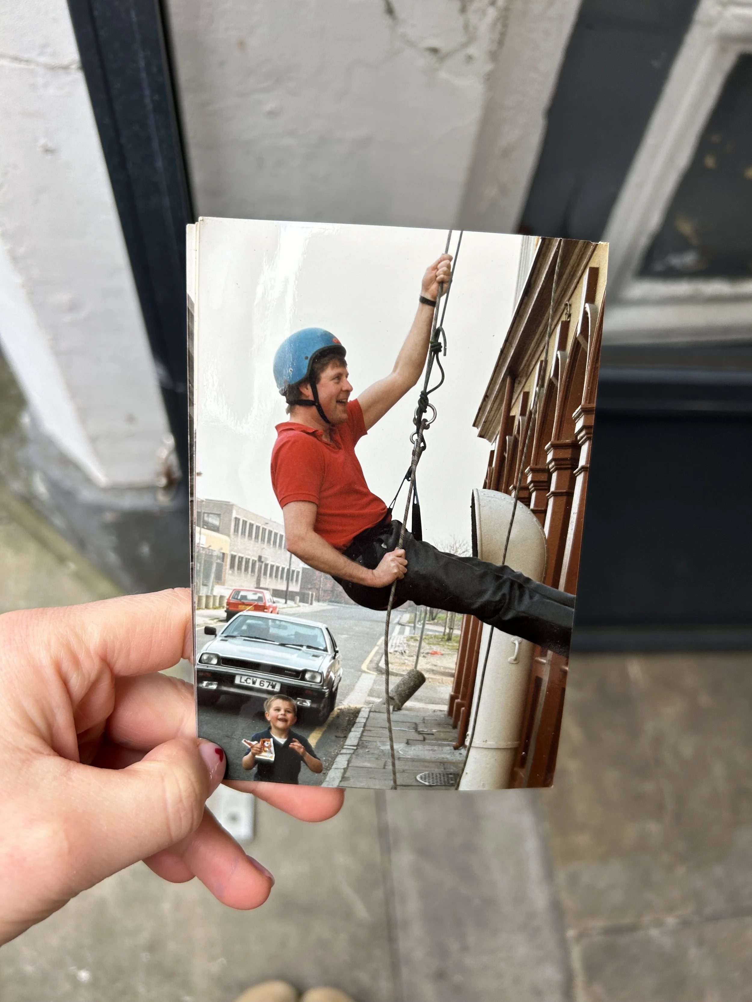 A person holding a photograph of a man wearing a blue helmet and red shirt, swinging on a rope, with a young boy standing on a street holding a toy airplane.