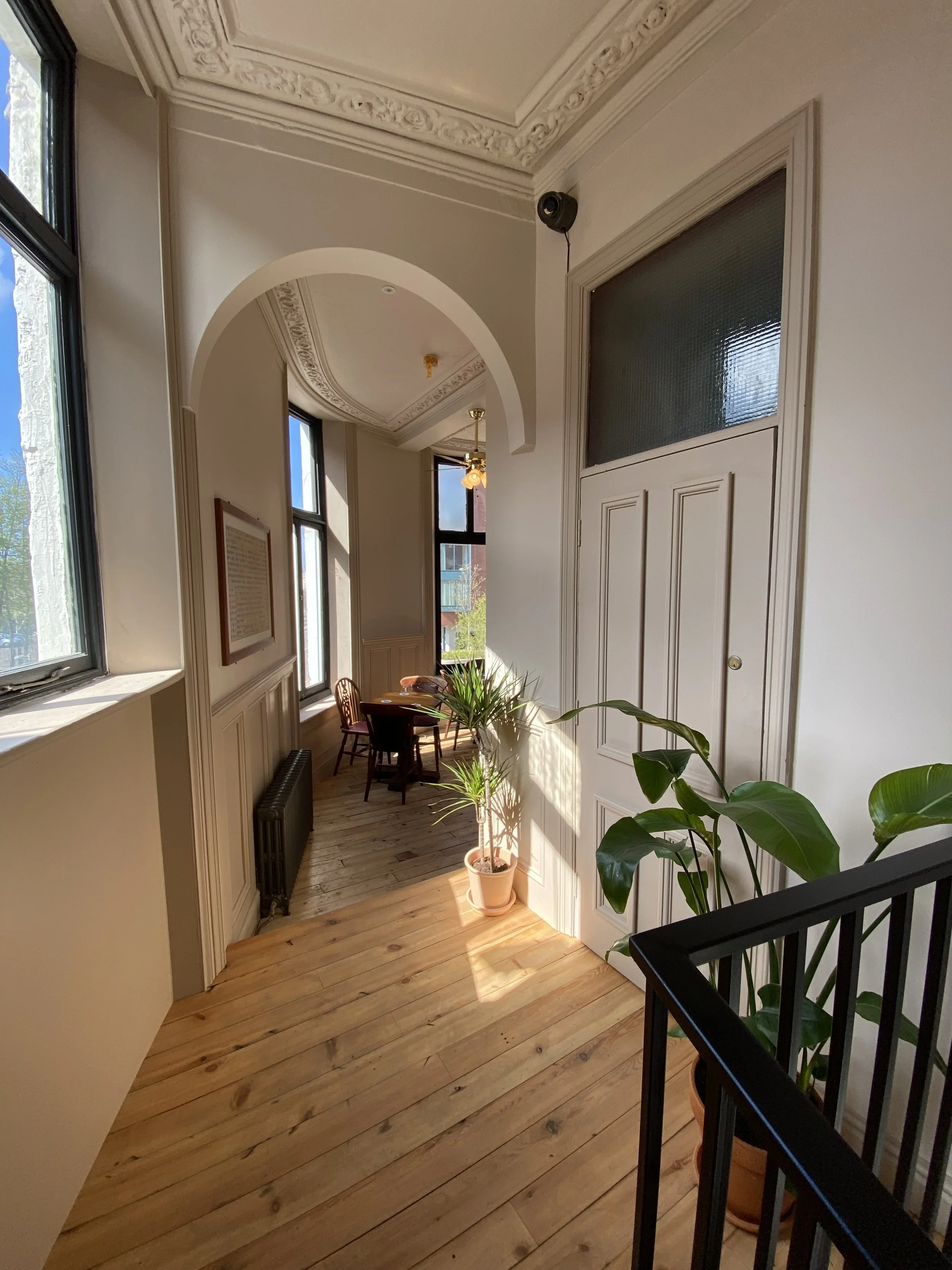Interior view of a hallway with wooden flooring, potted plants, and a window, leading to a small dining area with a round table and chairs. Decorative molding on the ceiling and walls, with natural light coming through large windows.