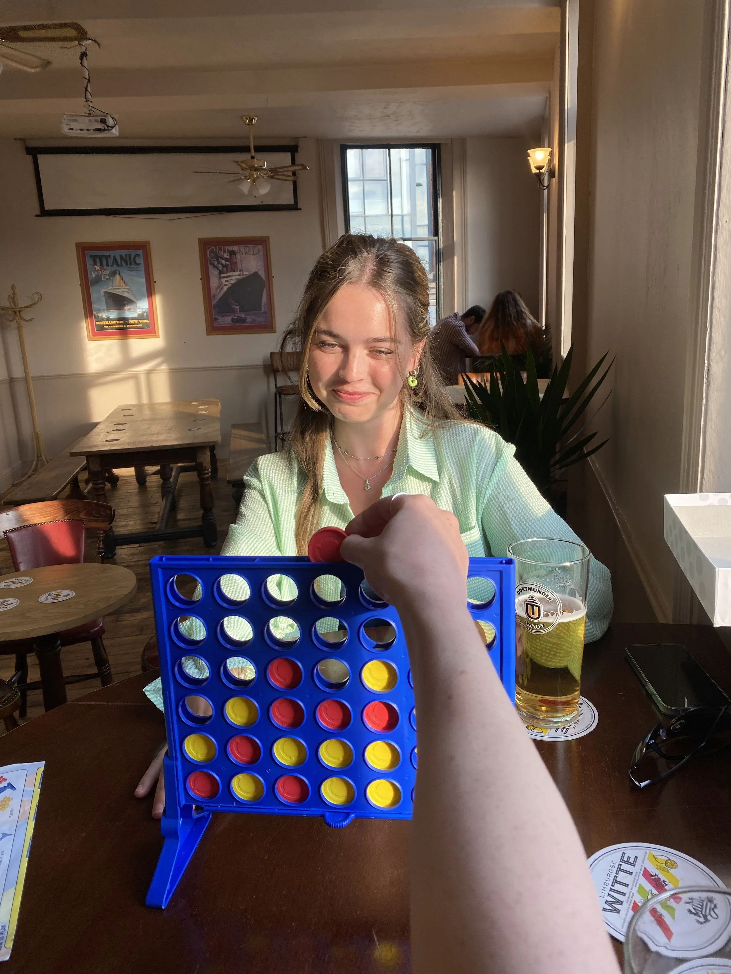A woman with long hair and earrings smiling as she plays Connect Four at a table in a sunny cafe or bar, with a glass of beer, sunglasses, and some menus on the table.