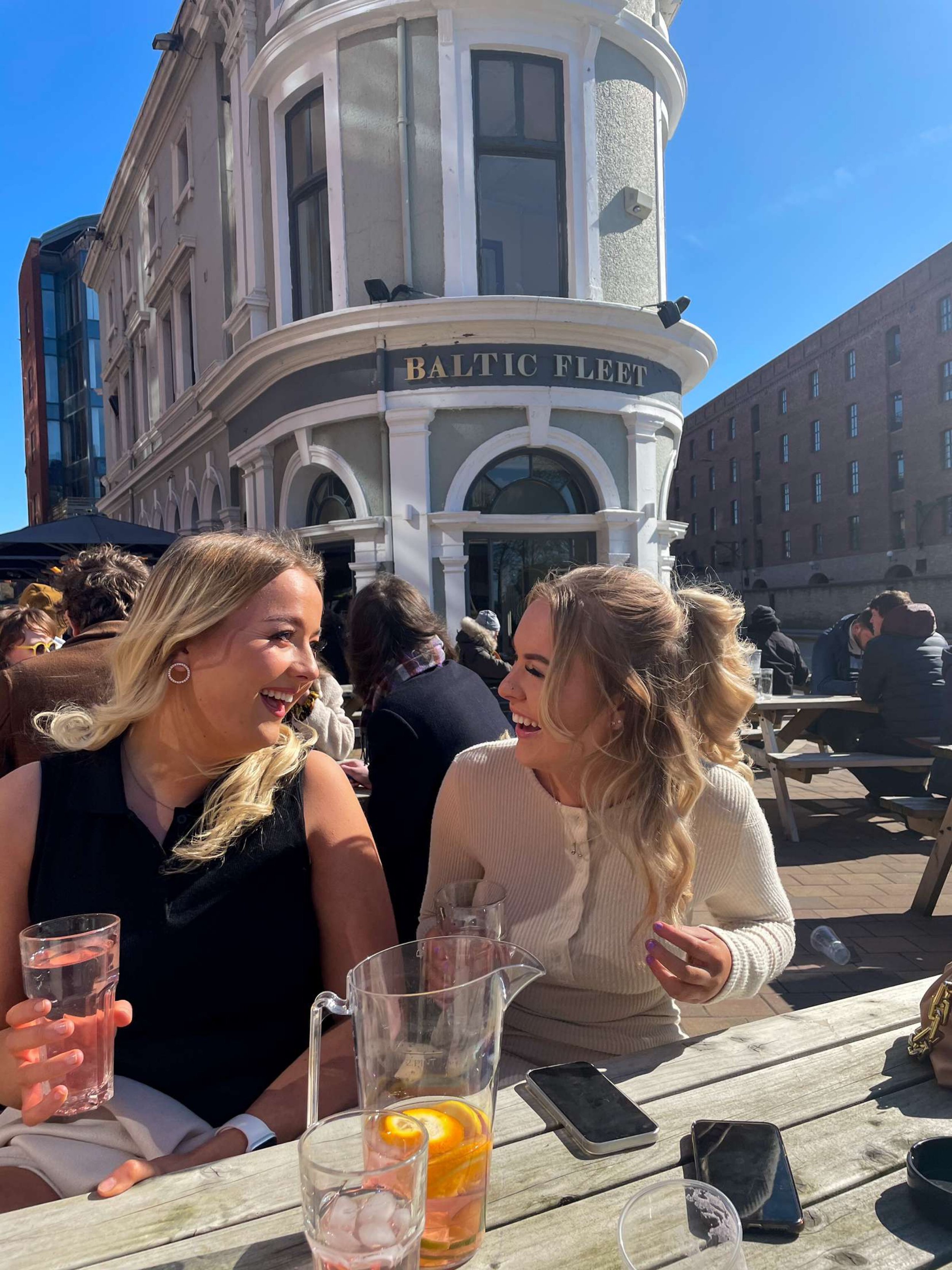 Two women sitting at an outdoor table, laughing and talking, with drinks and phones on the table, in front of a historic building labeled 'Baltic Fleet' on a sunny day.