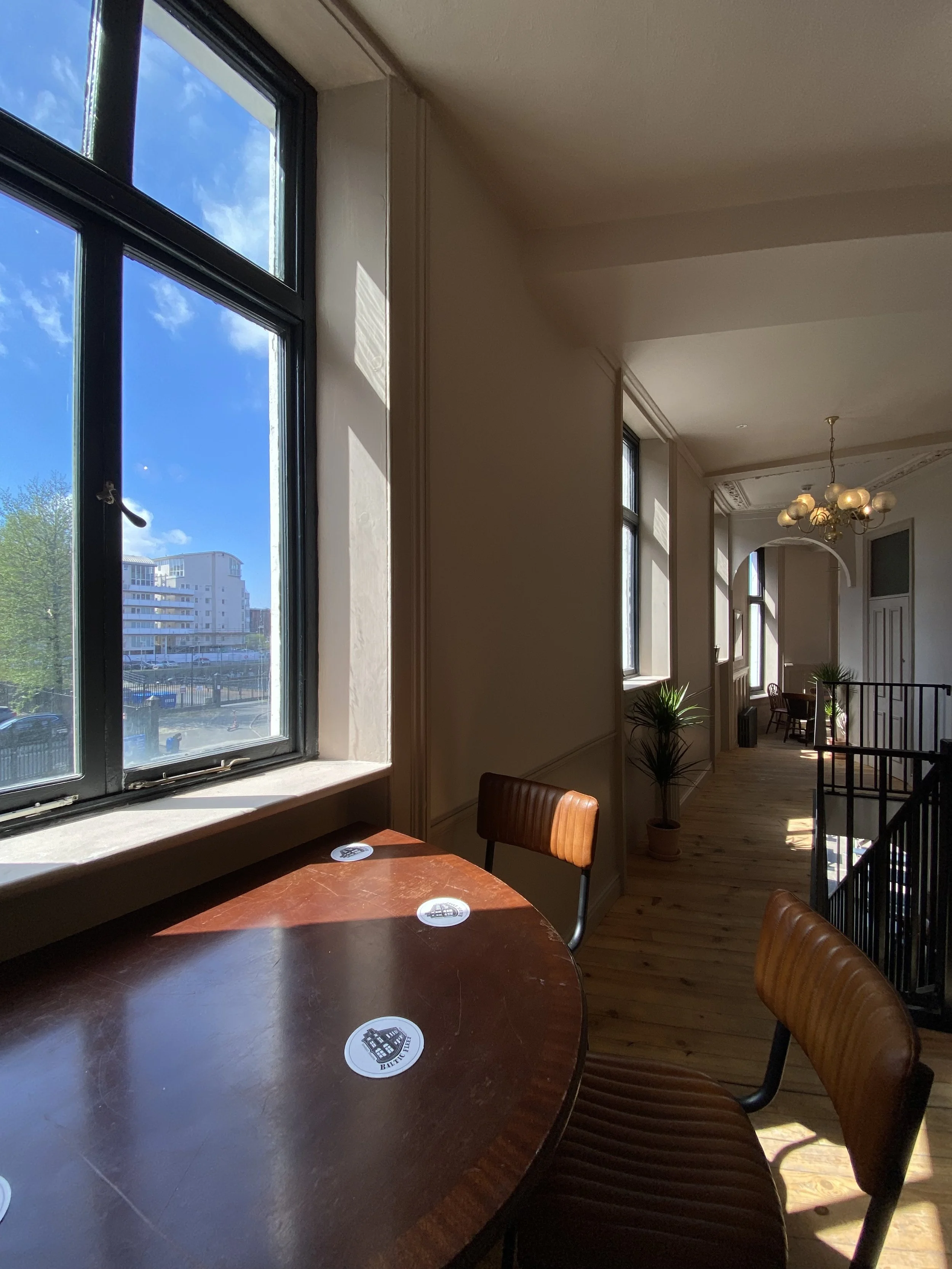 Interior of a cafe or restaurant with large windows, a round wooden table with chairs, sunlight casting shadows, and a chandelier hanging from the ceiling.