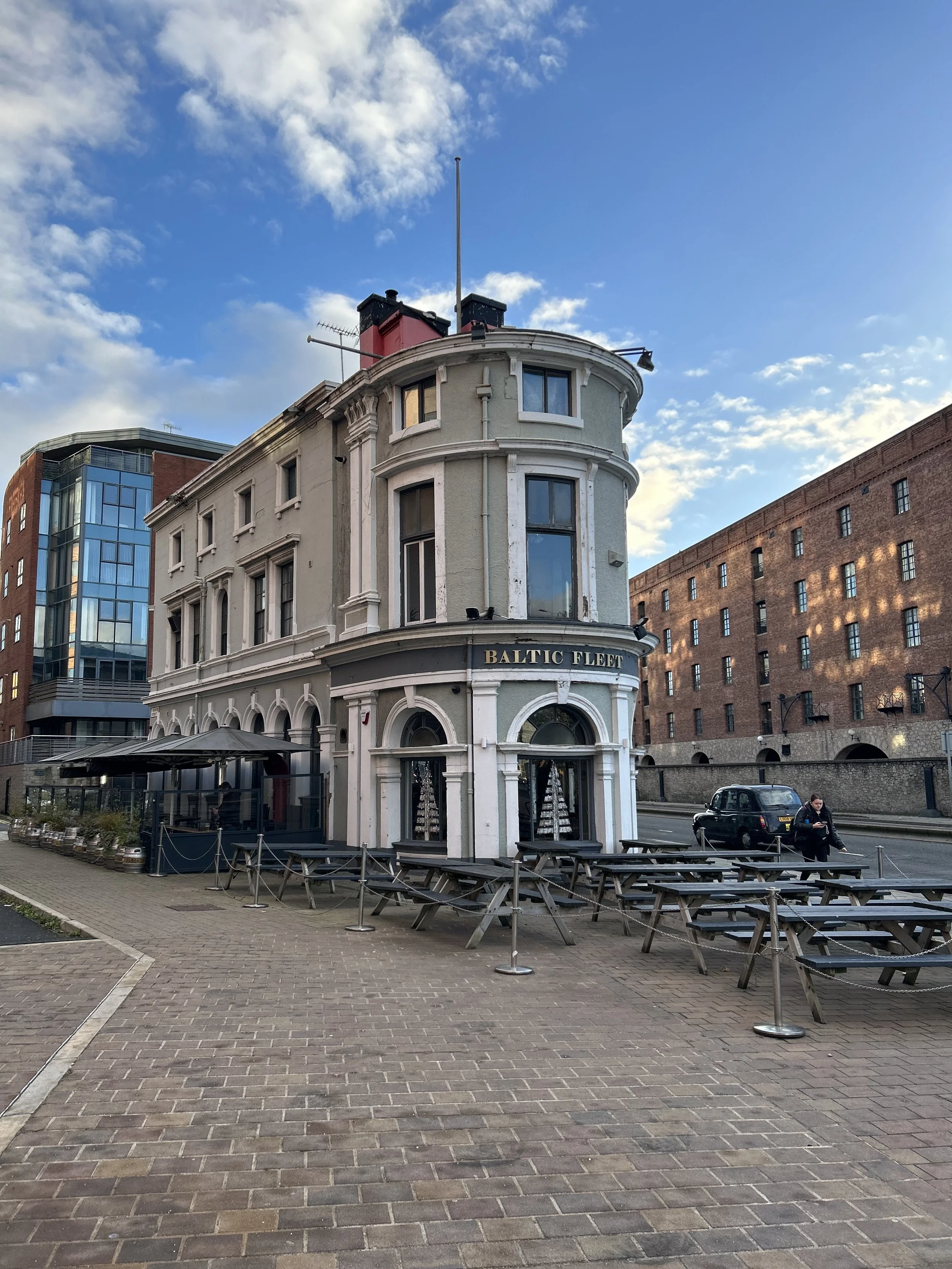 A three-story building with a sign that reads 'Baltic Fleet' on the upper floor, featuring large arched windows, situated in an urban area with modern buildings in the background. Outdoor seating with picnic tables and umbrellas is in front of the bu