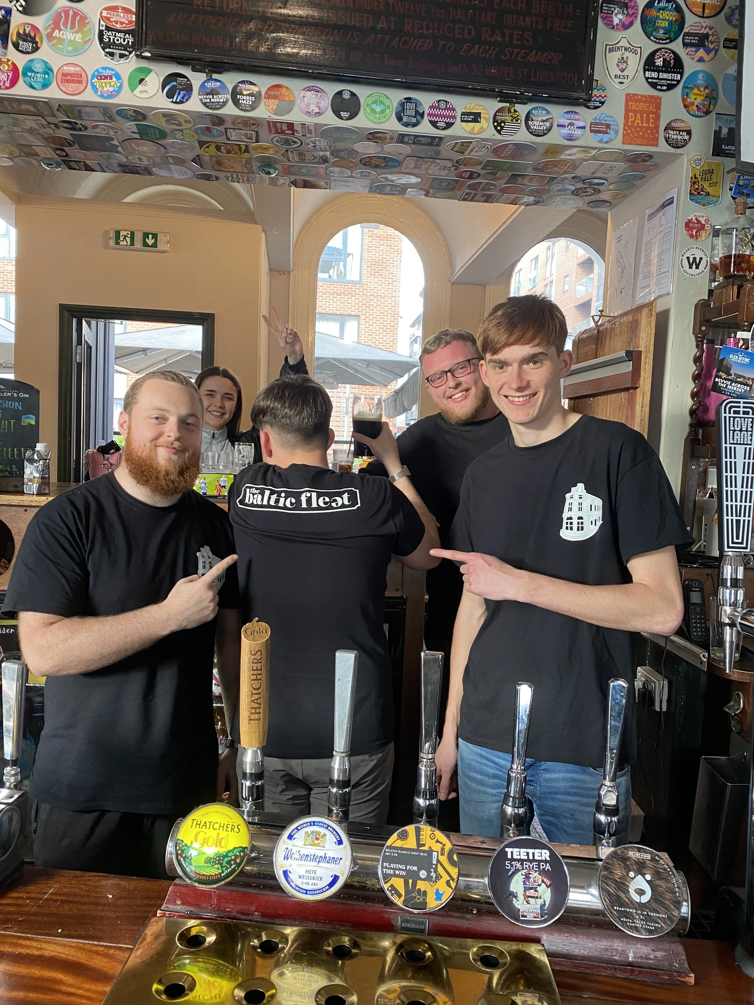 Four young men posing behind a bar with taps, with one person showing a peace sign and others pointing at him, in a pub with stickers on the ceiling and bar counter drinks