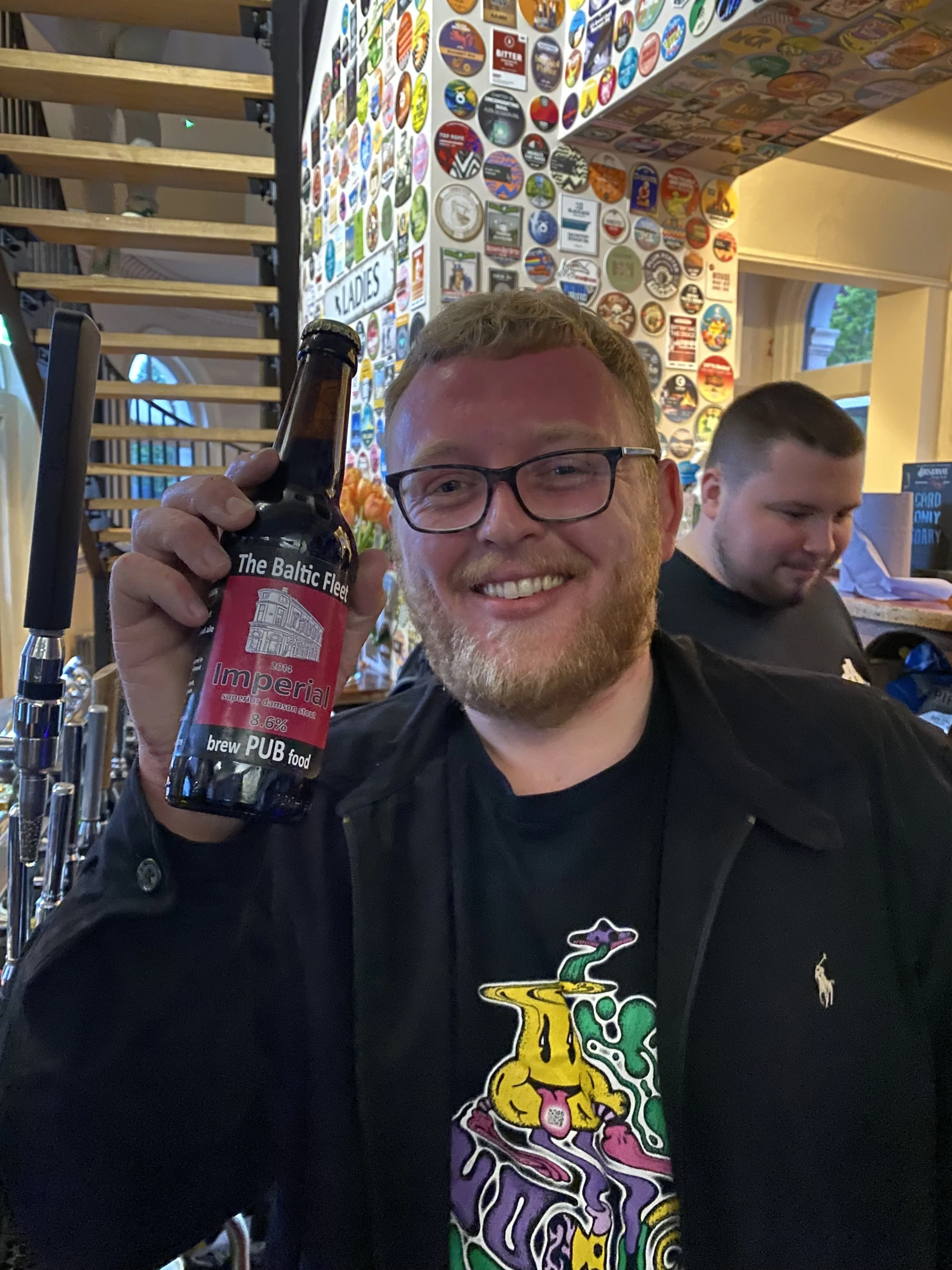 A smiling man with glasses holding a dark beer bottle inside a bar, with colorful wall decor and another person behind him.