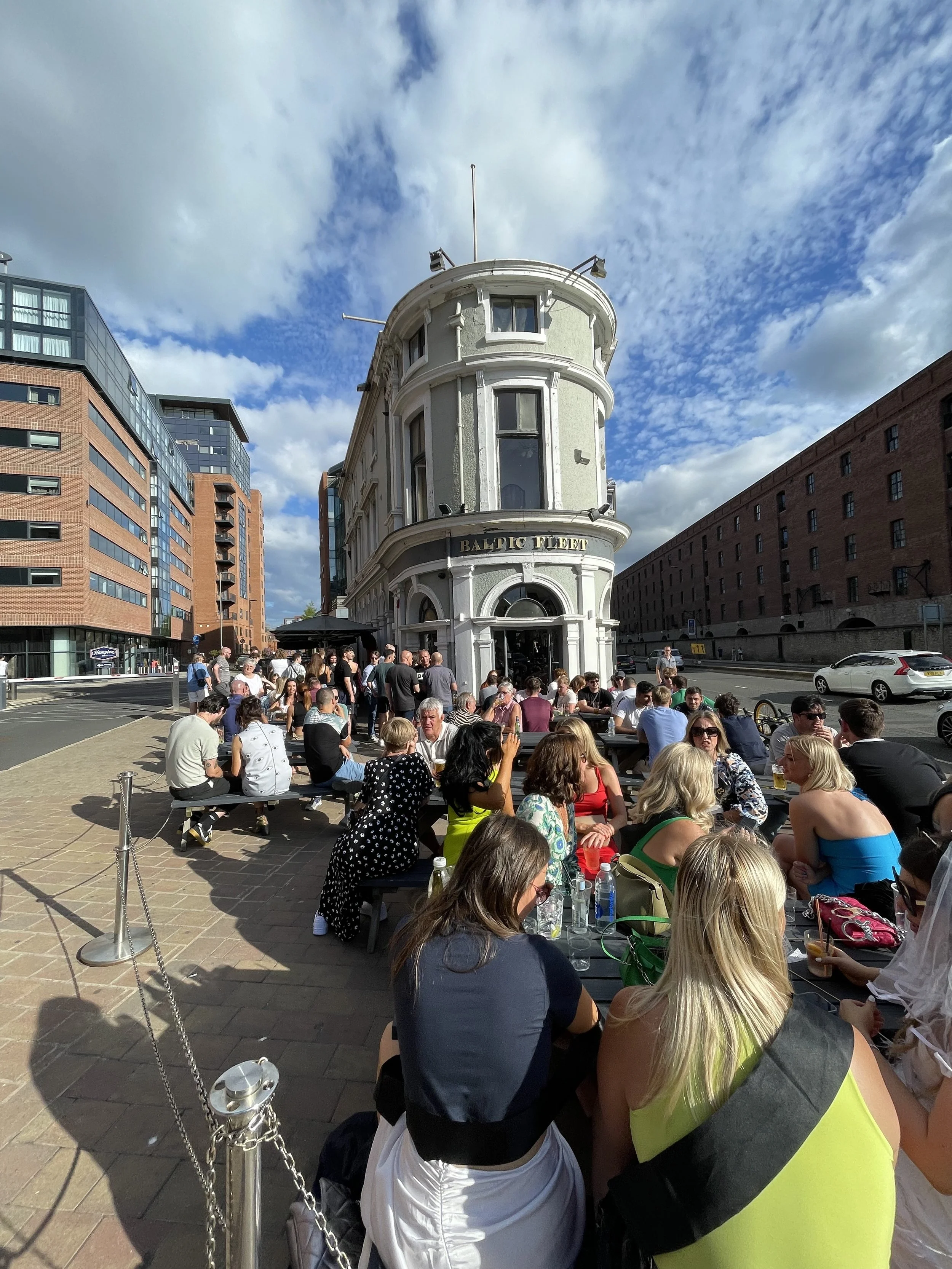 Crowd of people sitting and standing outside a white, rounded building labeled 'BALTIC FLEET' under a partly cloudy sky.