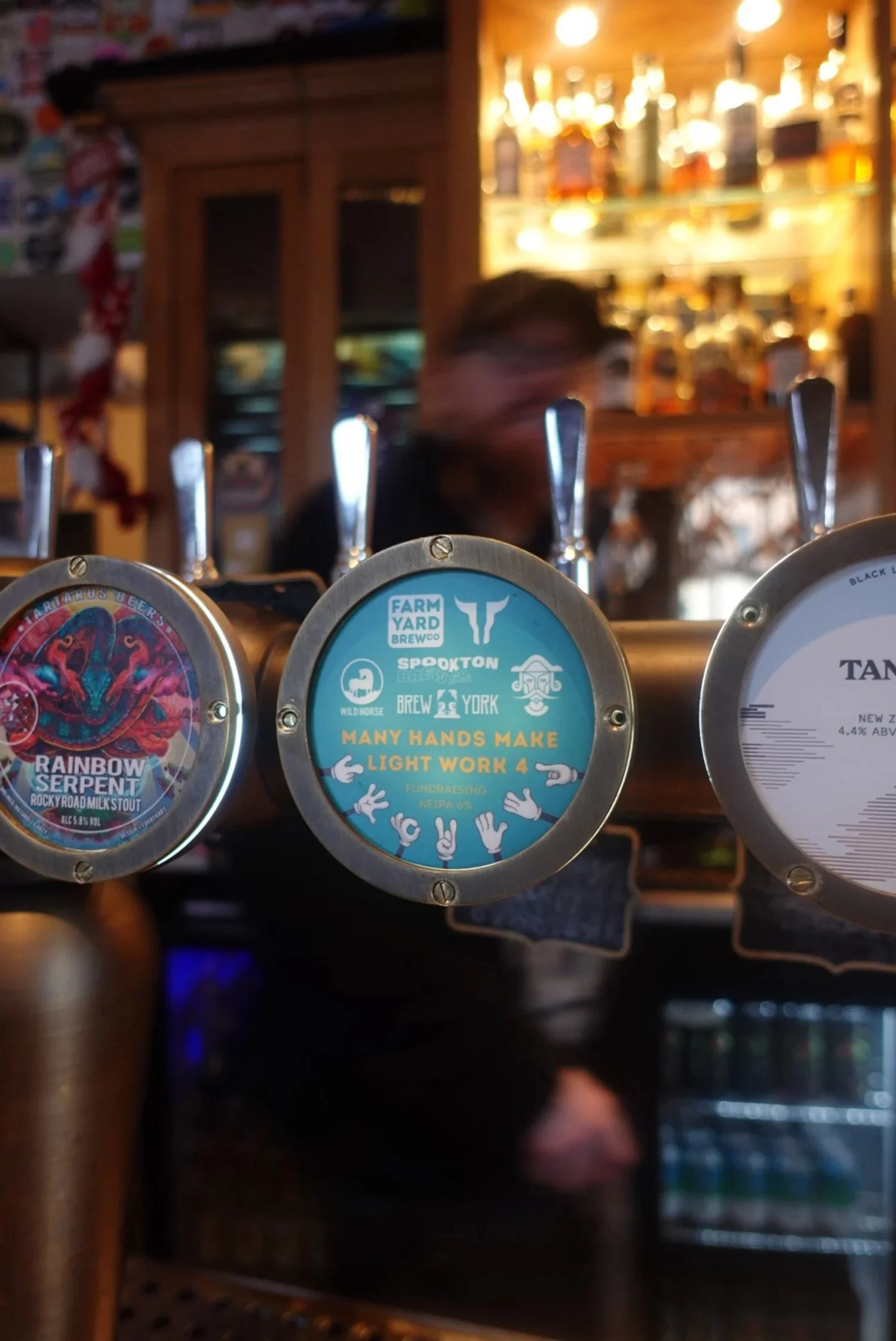 Close-up of beer tap handles, with the middle handle labeled 'Many Hands Make Light Work 4,' at a bar with a blurred bartender and liquor bottles in the background.